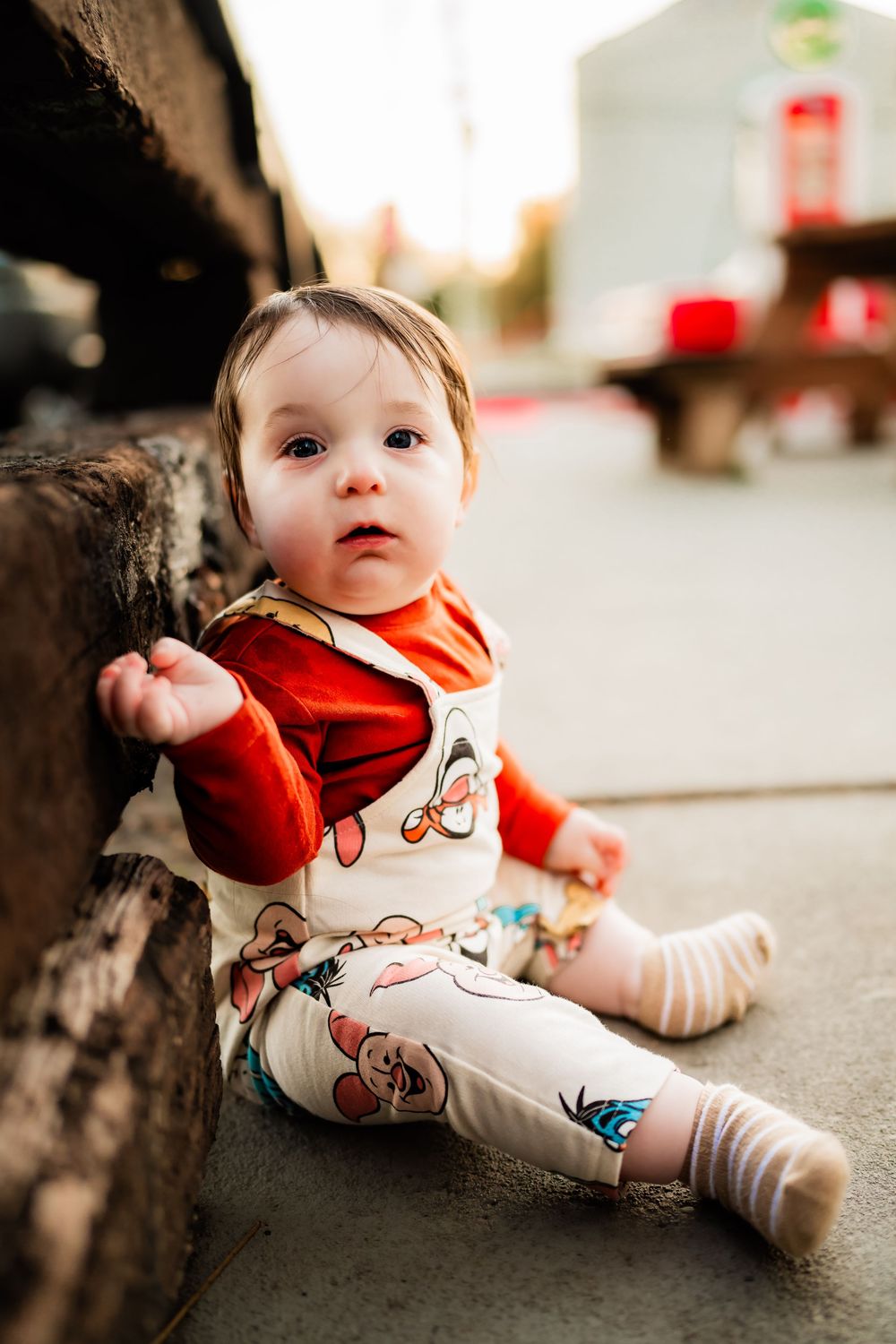 Adorable baby sitting on pavement leaning against wooden beam wearing red top and printed pants.