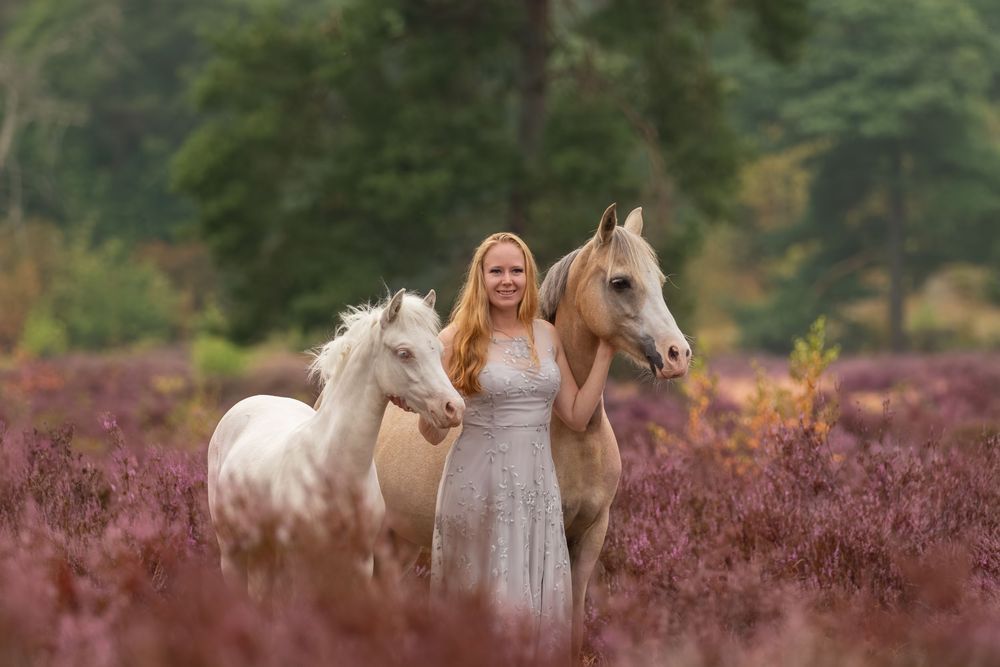 Glimlachende vrouw in een lange lichte jurk staat met een lichtbruin paard en een wit veulen tussen de bloeiende heide.