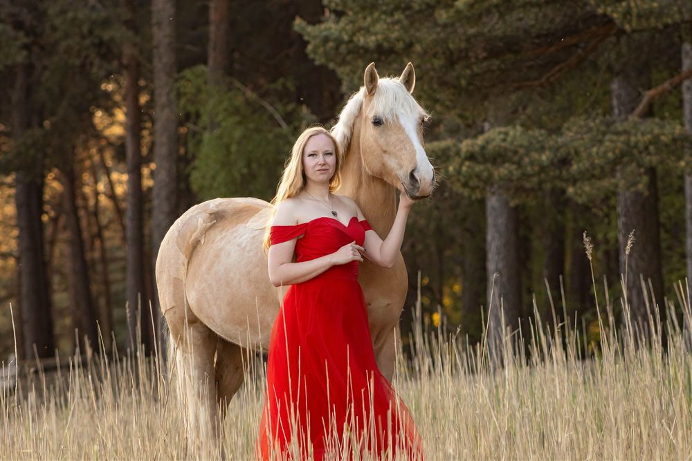 Vrouw in een lange rode jurk staat dicht tegen een lichtbruin paard aan in hoog gras bij het bos.