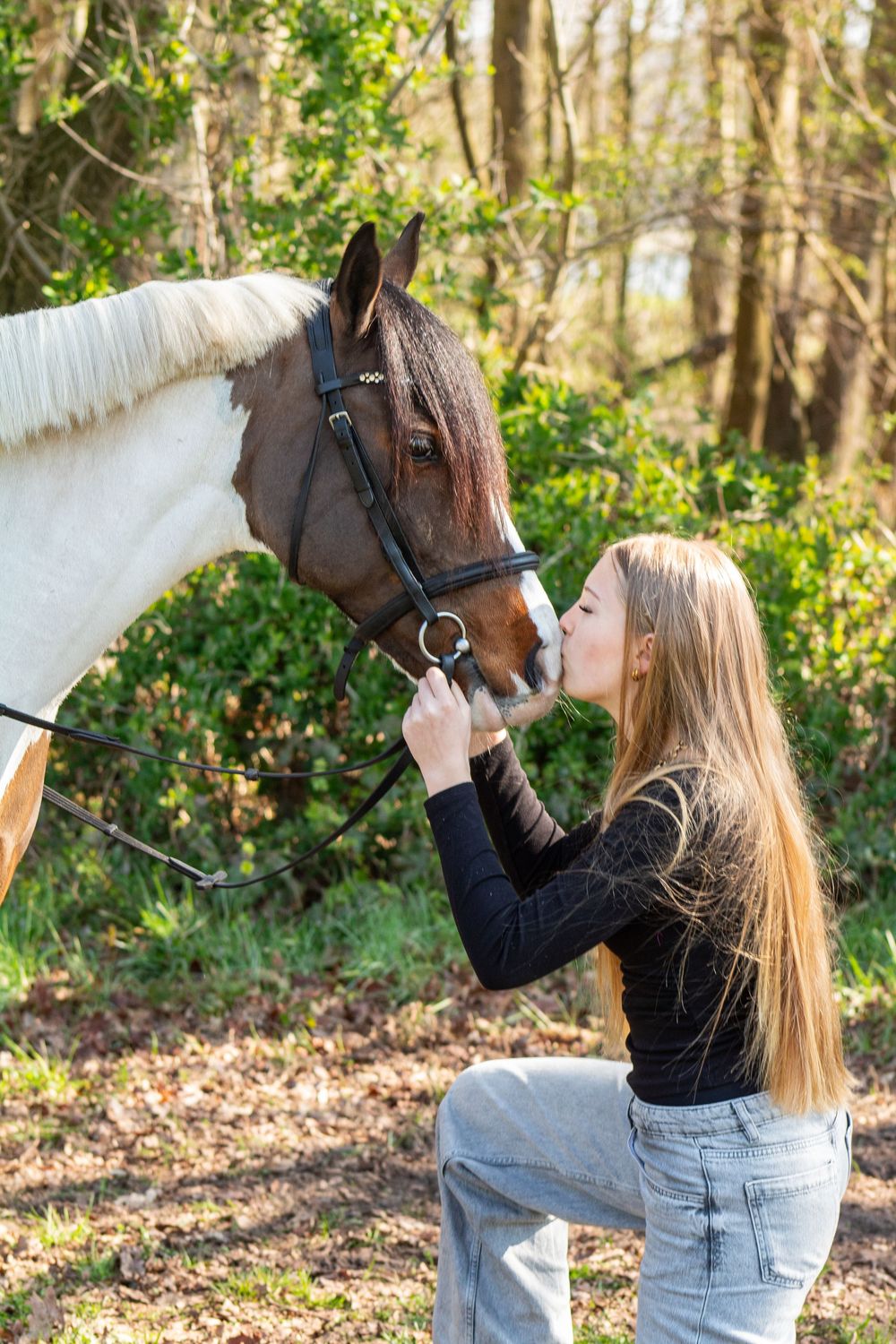 Jonge vrouw knielt in het bos en aait de neus van een bruin-wit gevlekt paard.