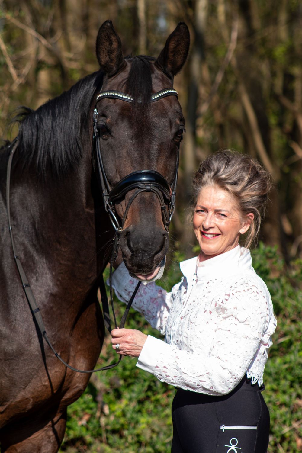 Glimlachende vrouw omhelst een zwart paard met een hoofdstel in een bosrijke omgeving.