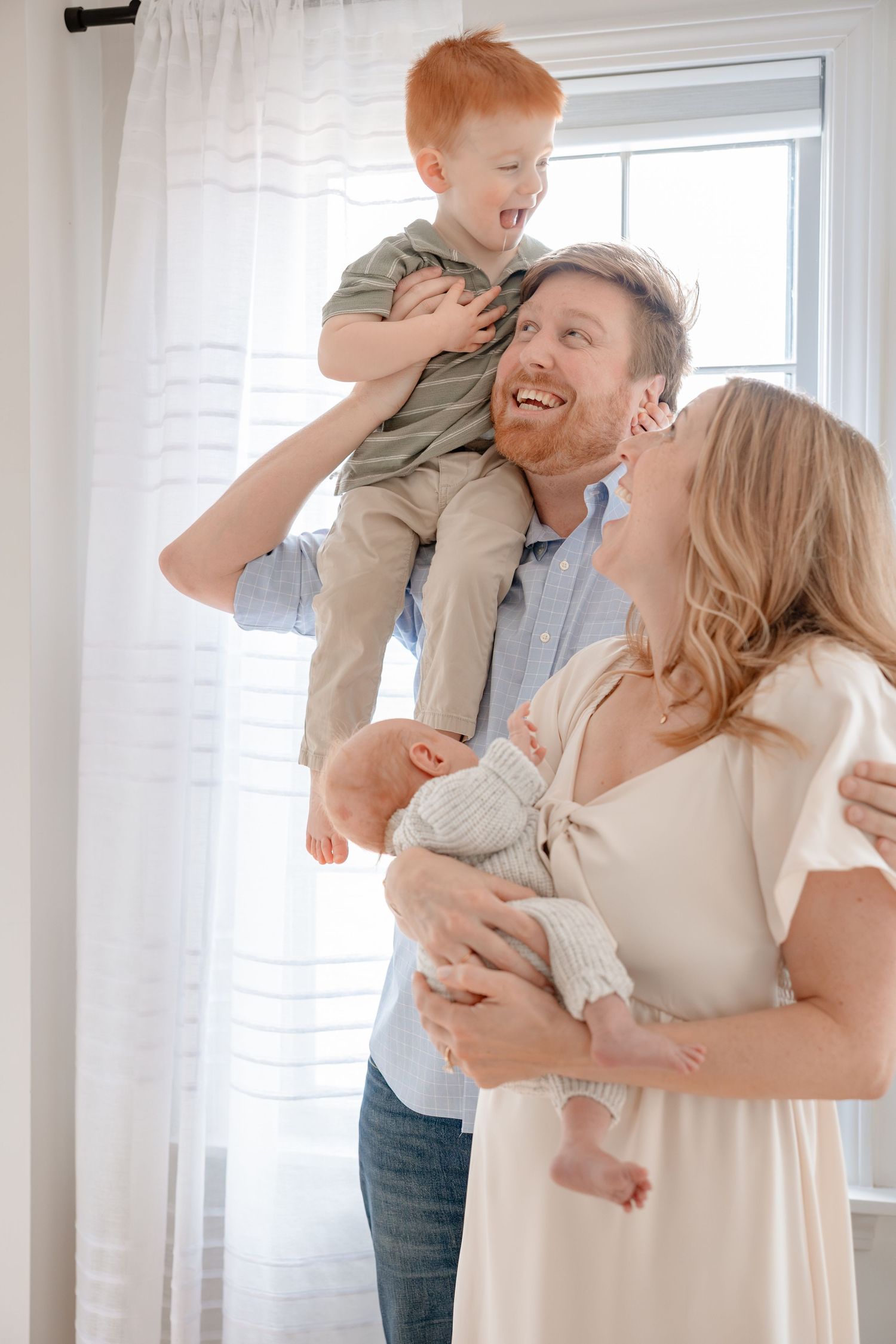 A couple in white clothing shares joyful moments with their baby in a bright, sunlit room.