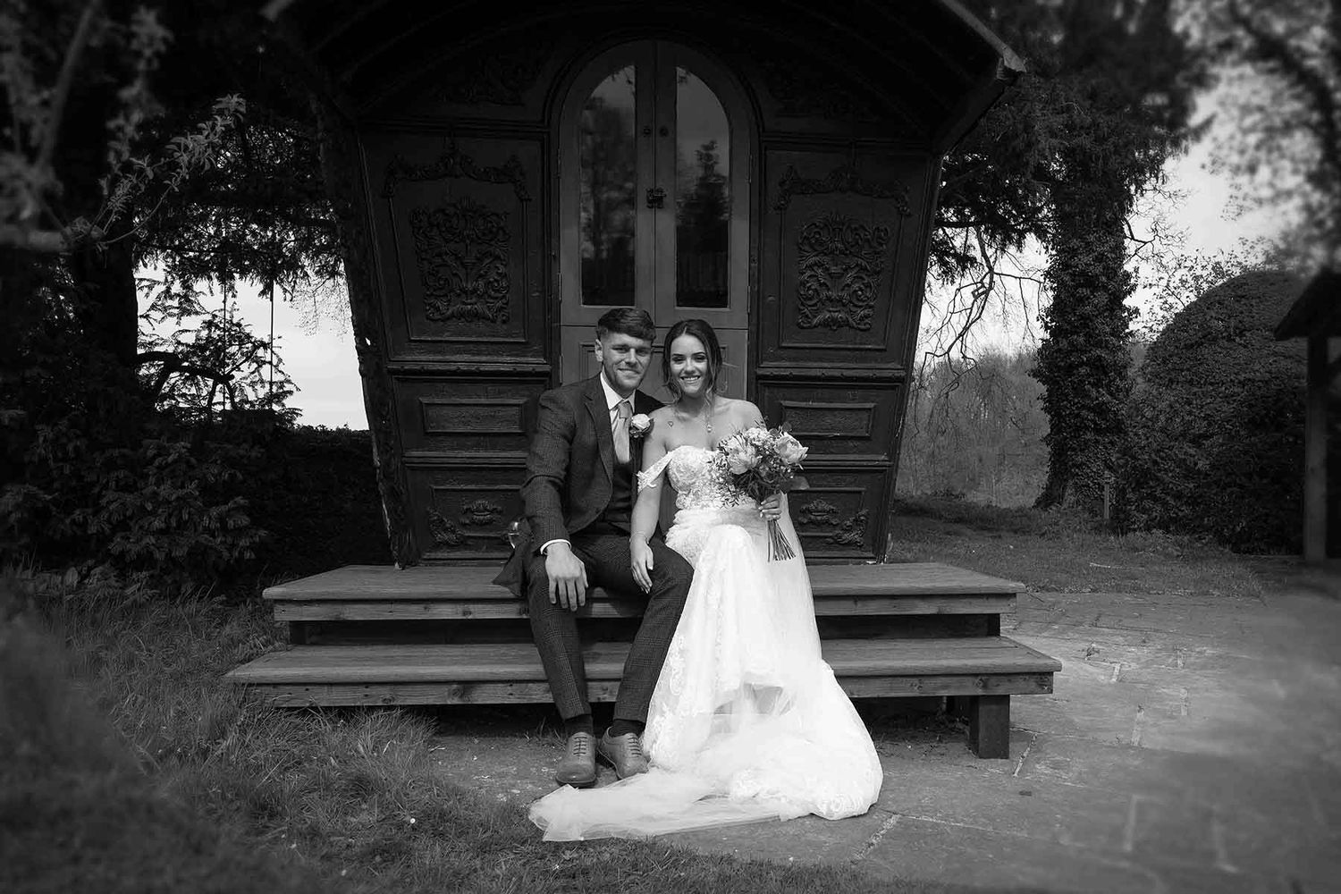 A black and white wedding photo of a couple posing together on wooden steps in front of a rustic chapel in the grounds of Askham Hall