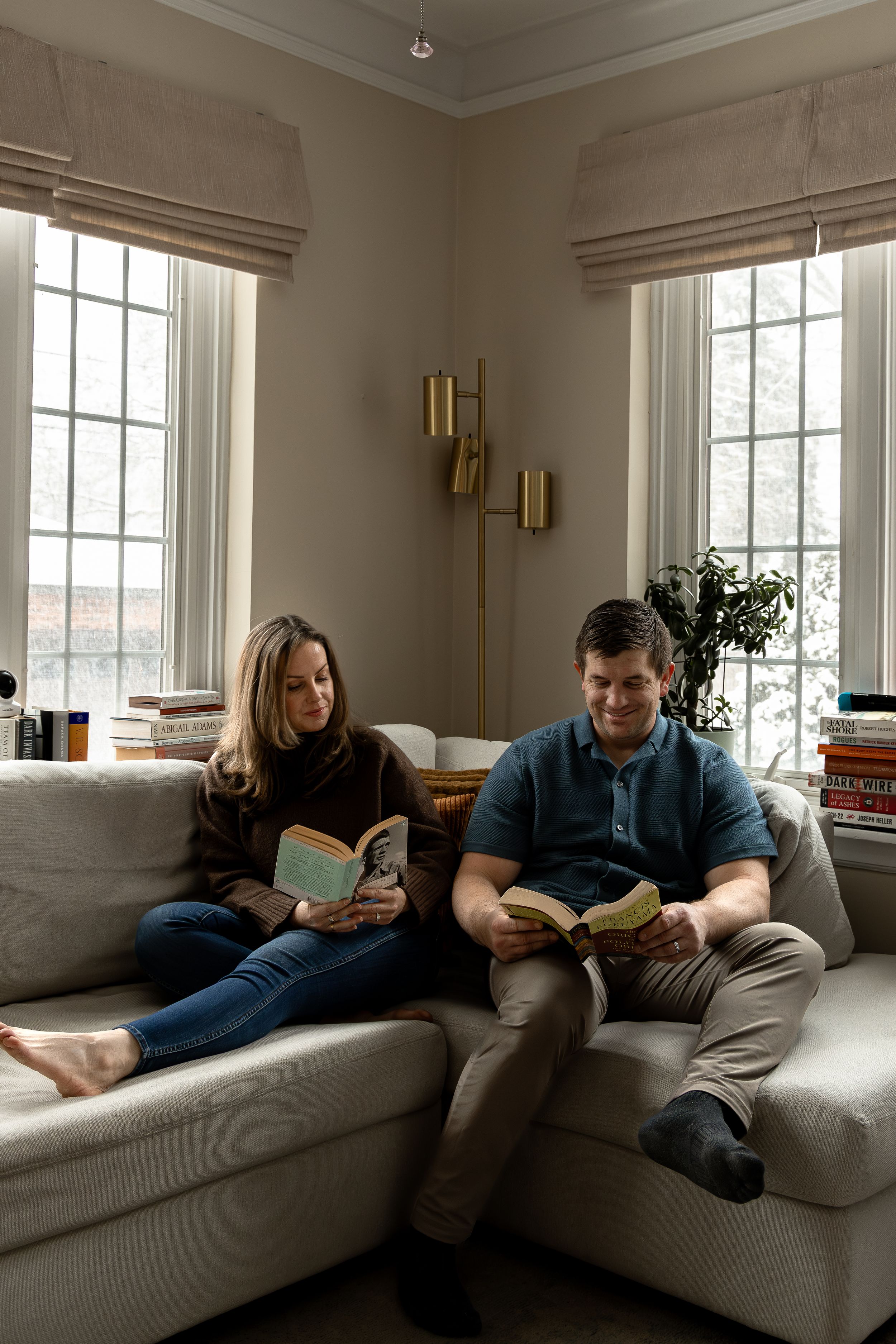 A couple relaxes on a couch in a bright room with large windows while reading books together.
