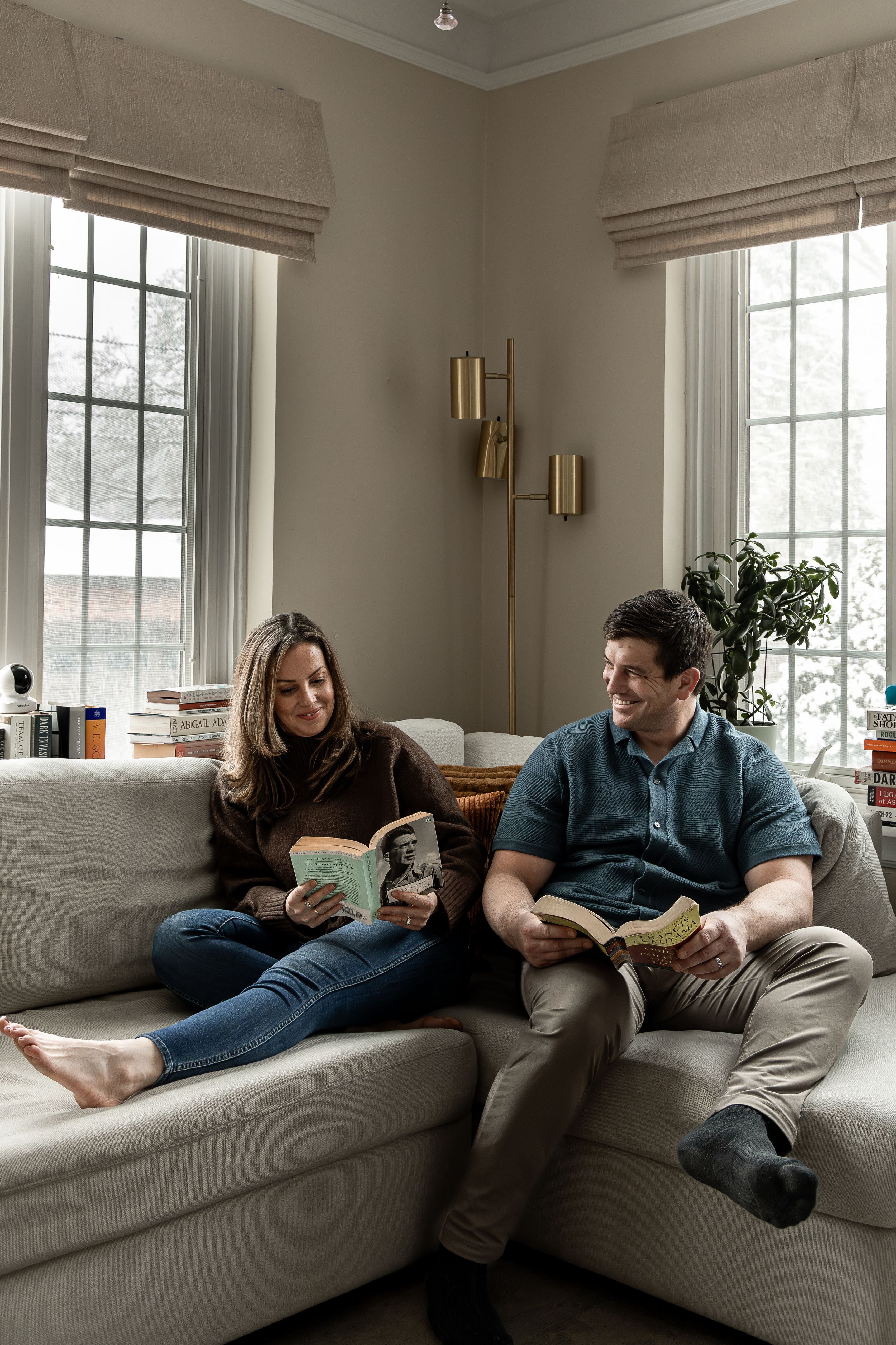 Two people relax on a gray couch by large windows while reading books together in a cozy living room.