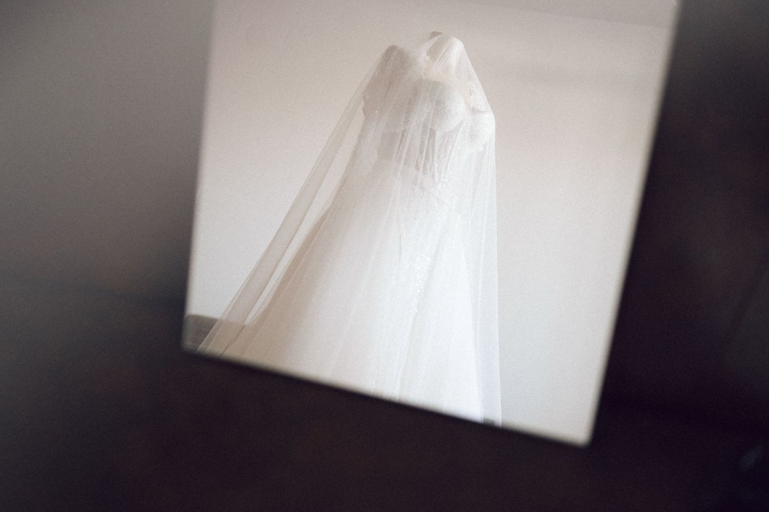 A white lace dress hangs from a wooden hanger against a white wall, creating an elegant bridal display.