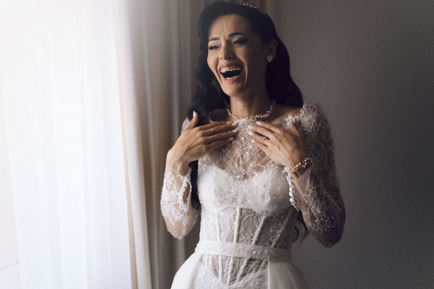 A bride in a white lace wedding dress laughs joyfully while standing near a window in natural light.