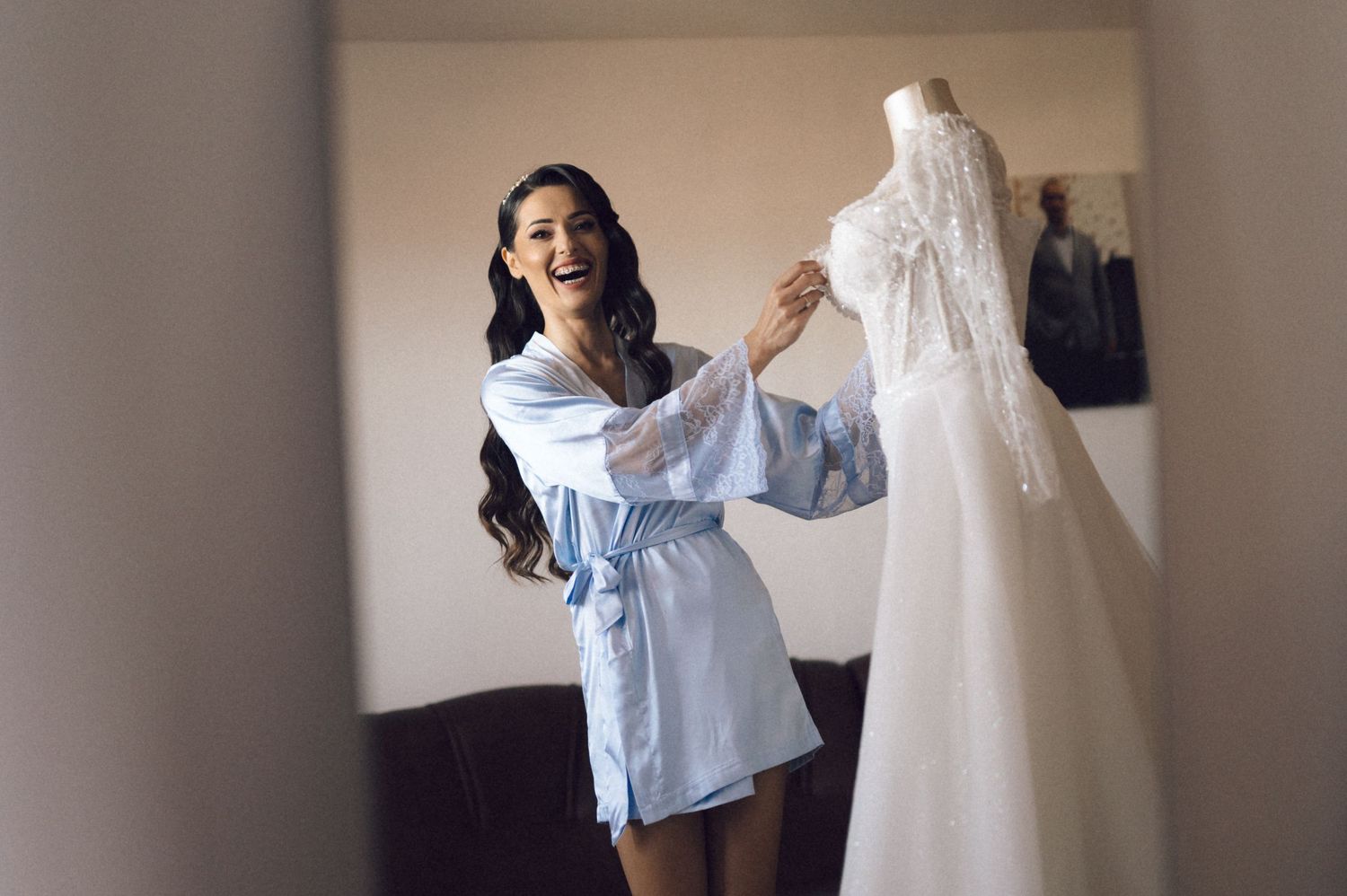 A woman in a blue robe stands excitedly next to a white wedding dress on display.
