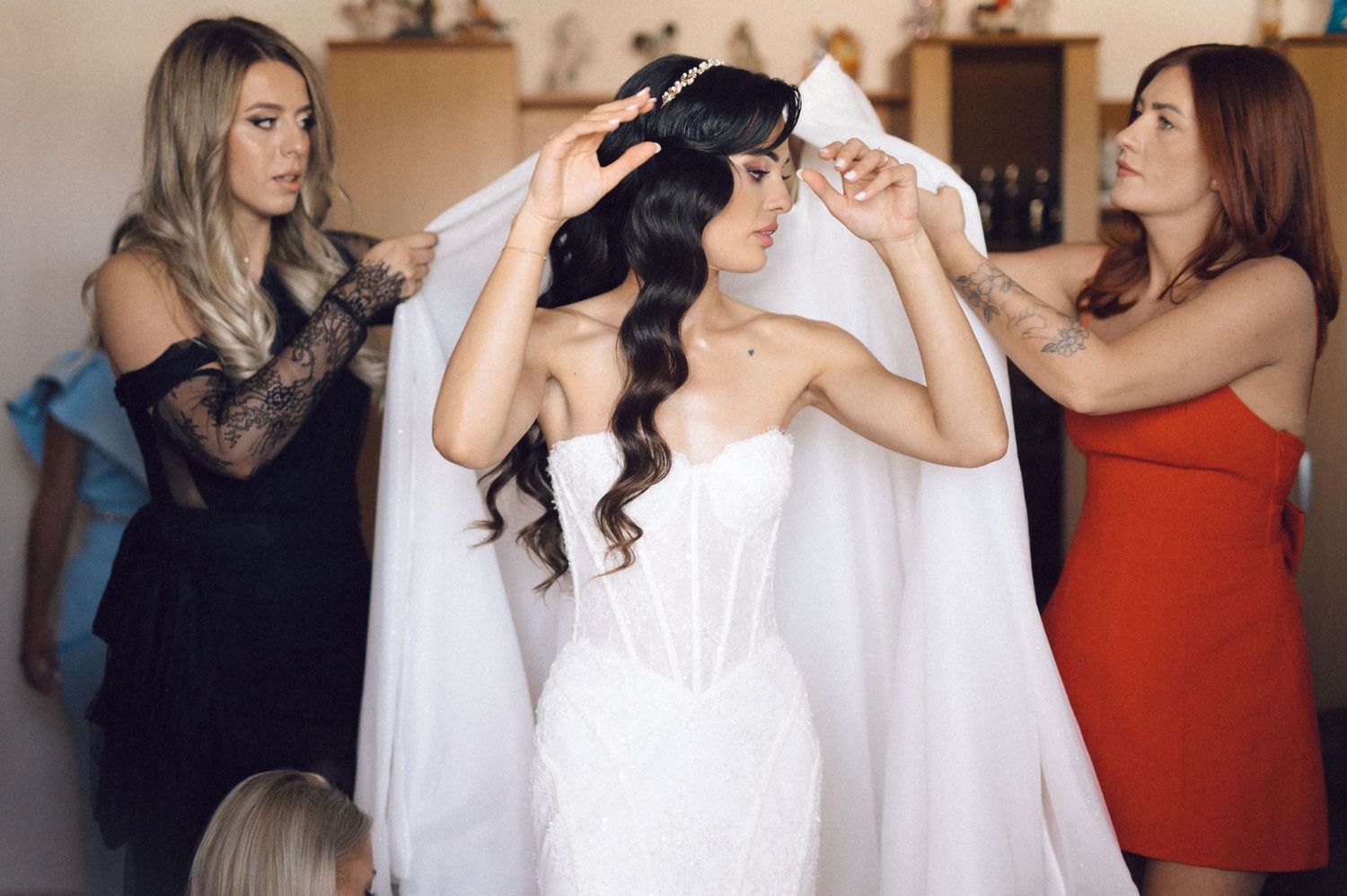 Bridesmaids help adjust a flowing white veil on a bride wearing a strapless wedding gown during pre-ceremony preparations.