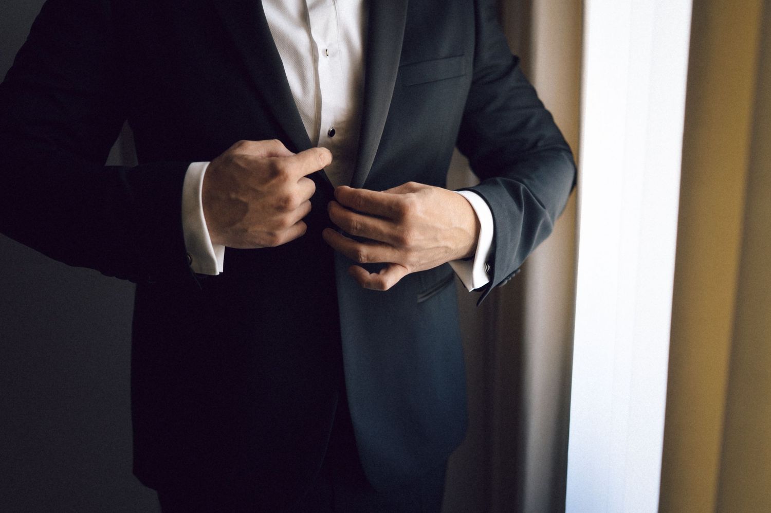 A person in a dark suit adjusts their tie while standing in front of a mirror.