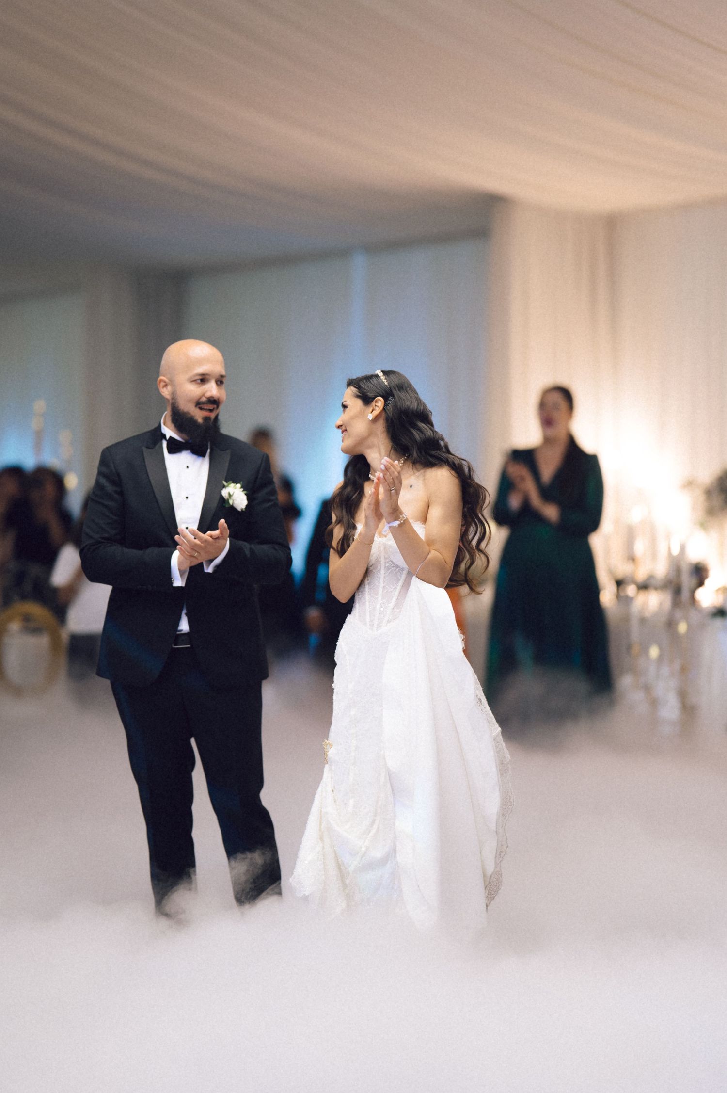 Newlyweds share their first dance together in a white reception hall filled with fog and guests watching.