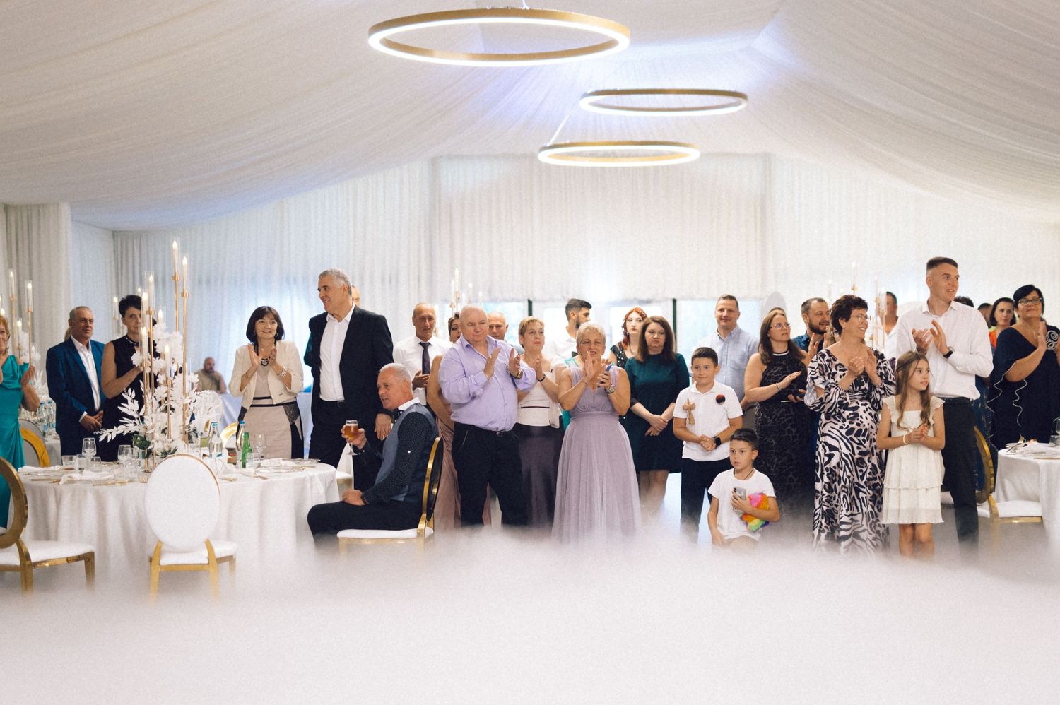 Wedding reception guests stand and applaud in an elegant white room with circular pendant lights overhead.