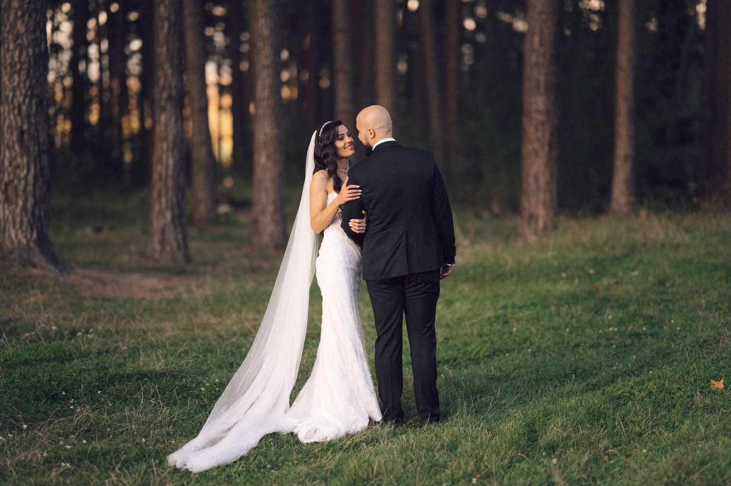 A bride and groom share an intimate moment in a sunlit forest setting with a flowing white wedding veil.