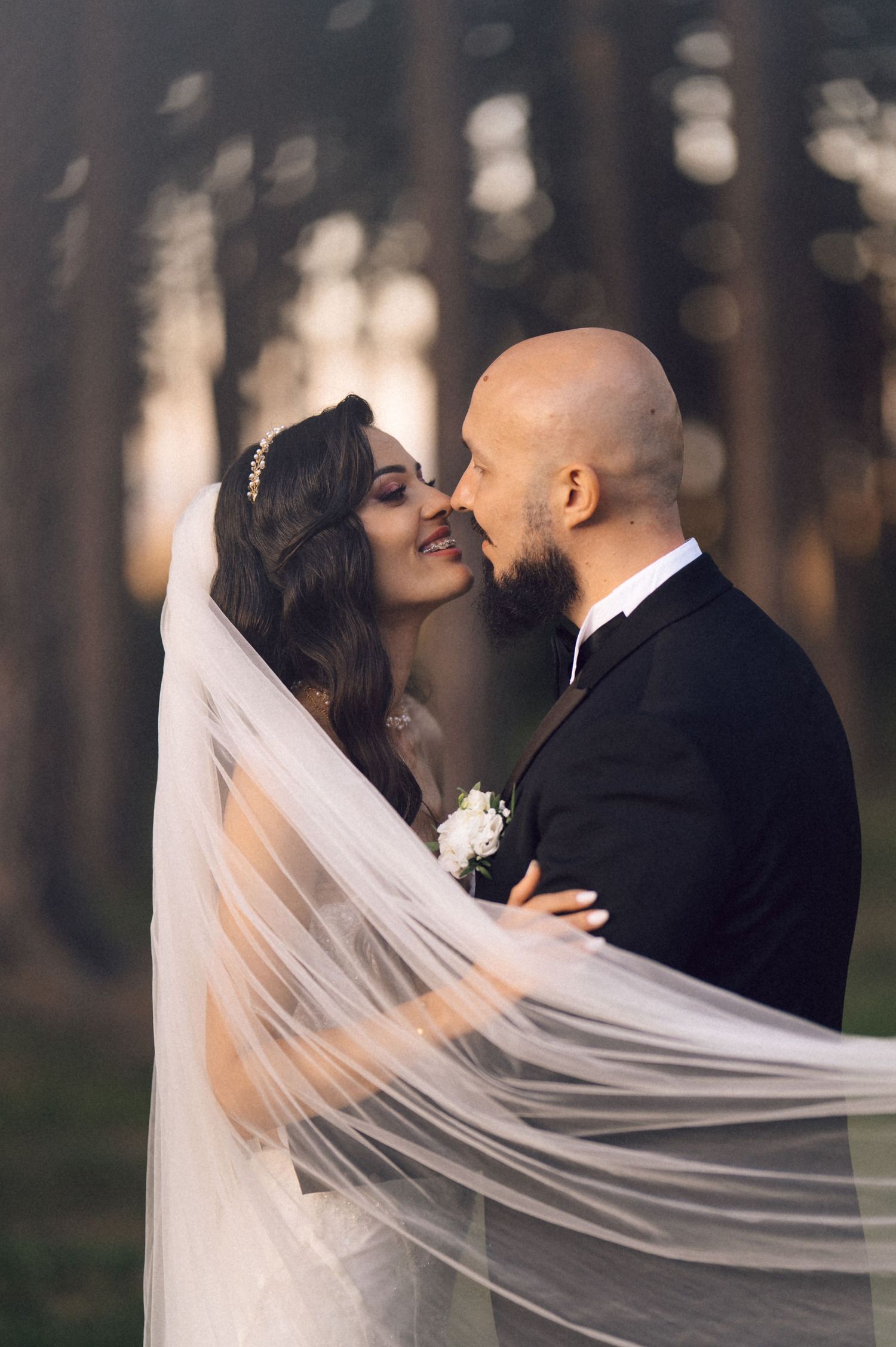 A romantic wedding couple shares an intimate kiss in a forest setting as the bride's long veil flows in the breeze.
