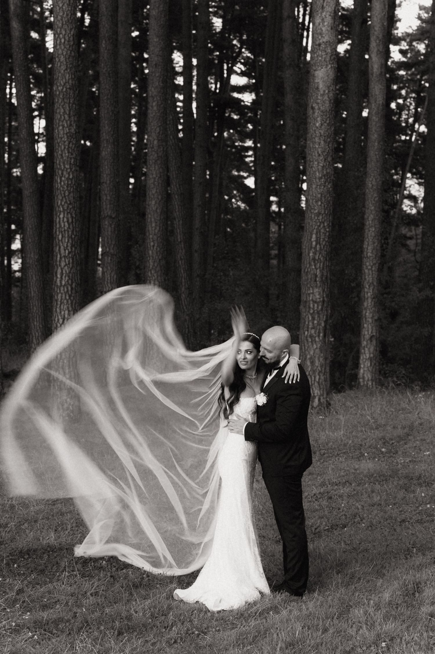 A wedding couple stands in a forest as the bride's veil flows dramatically in the wind creating an ethereal sequence.