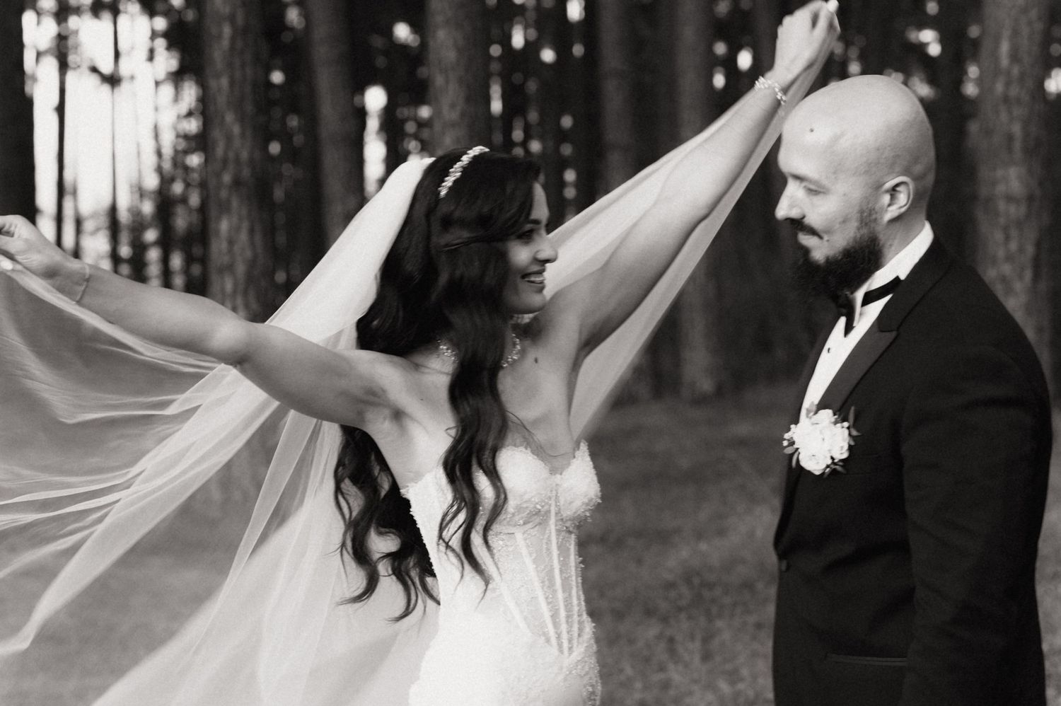 A joyful black and white wedding photo captures a playful moment with a flowing veil in a serene forest setting.