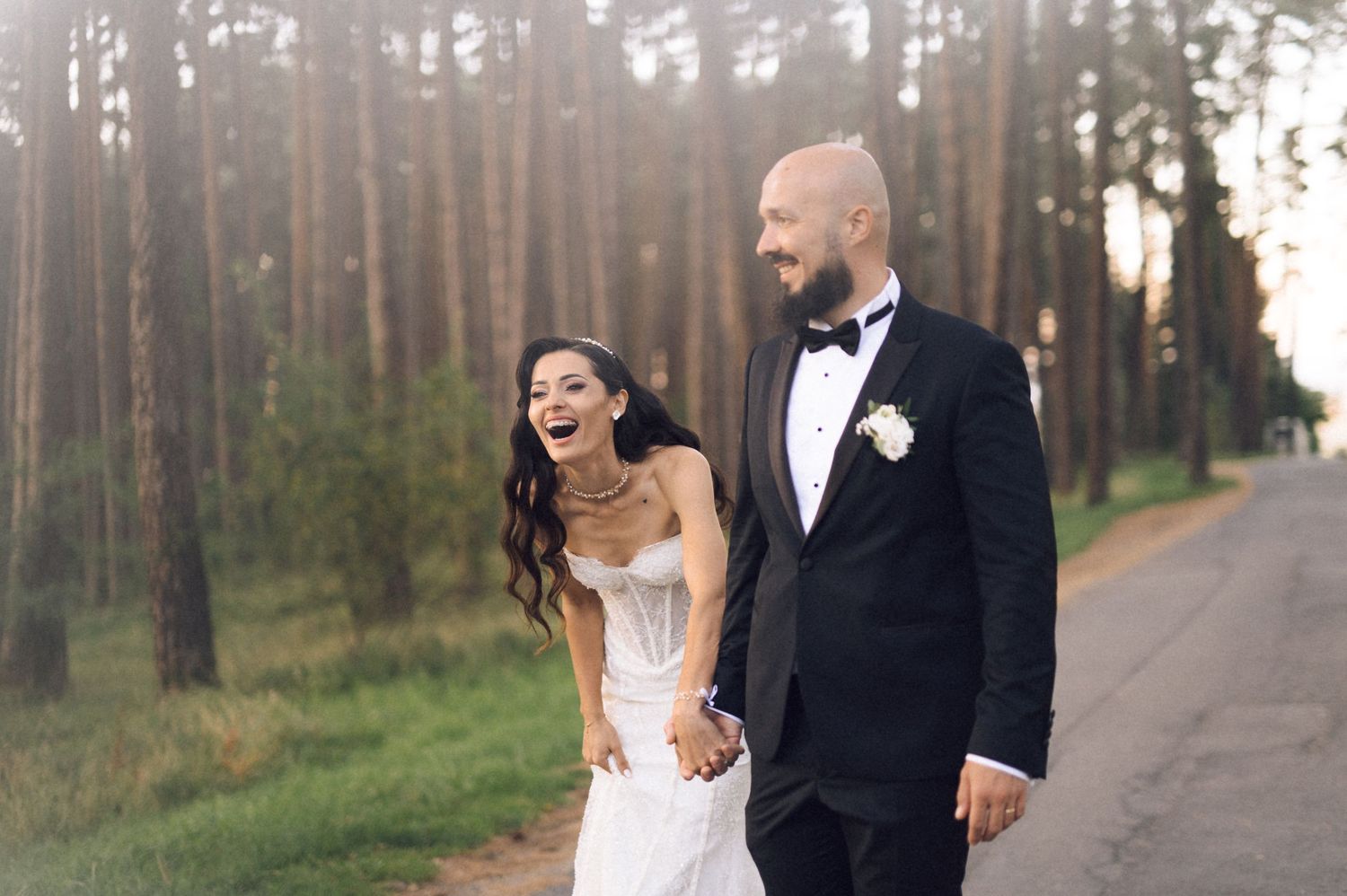 Couple walking hand in hand on a tree-lined path wearing formal wedding attire during sunset.