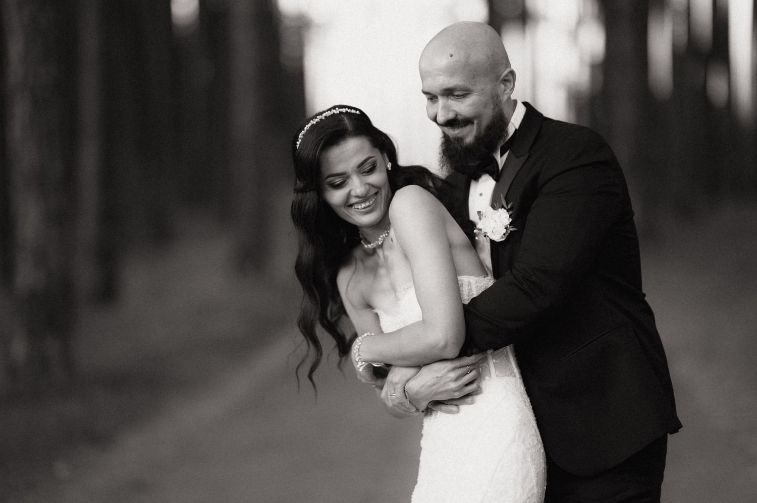 A romantic black and white wedding photo shows a couple embracing and laughing together in a wooded setting.