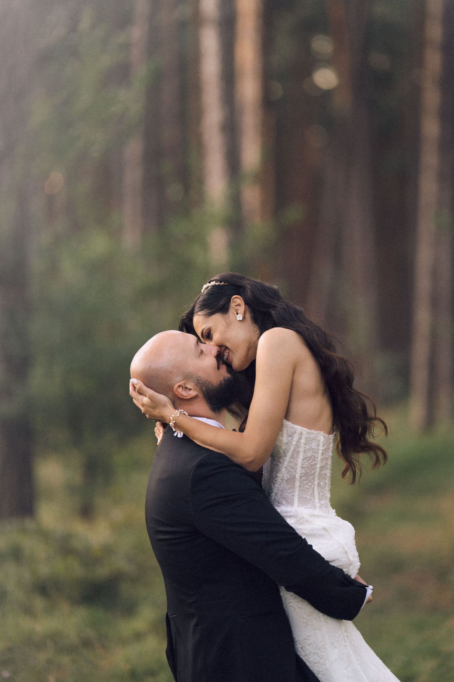 A romantic wedding couple embraces in a serene forest setting with tall pine trees in the background.