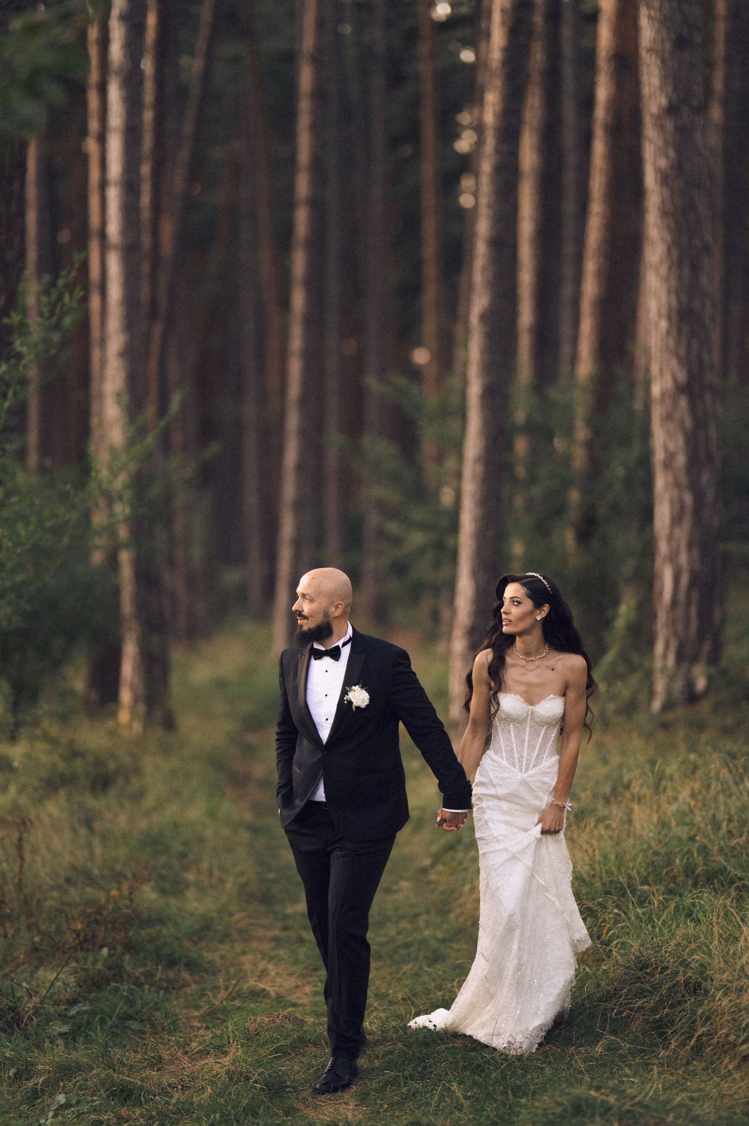 A couple in wedding attire walk hand in hand through a serene pine forest at dusk.
