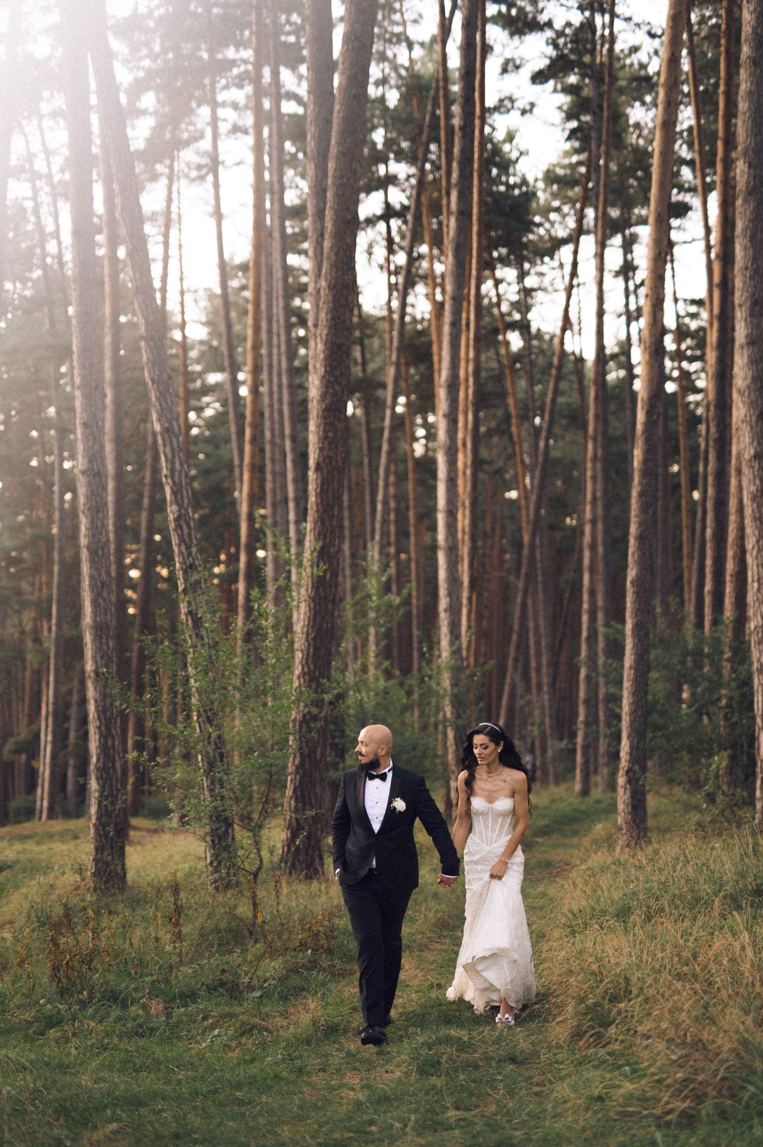 A couple in wedding attire walks hand in hand through a serene pine forest at sunset.