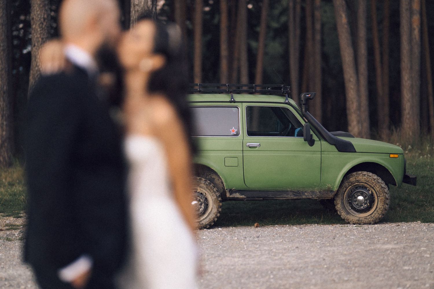 A couple embraces during their wedding photo session in a wooded area with a vintage green car in the background.