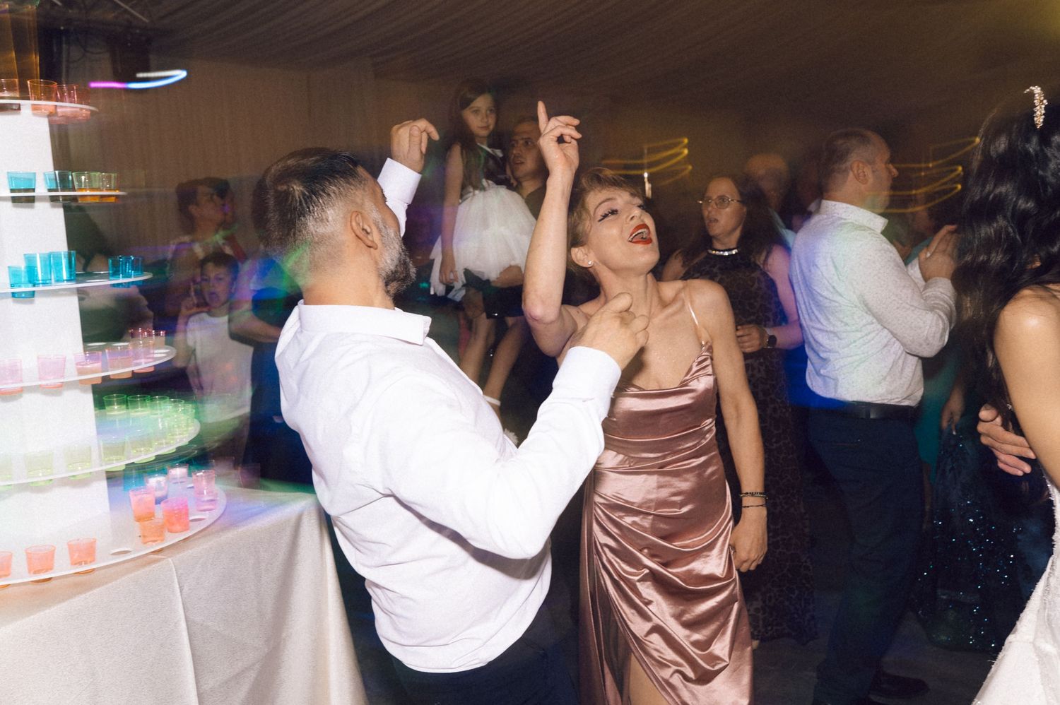 People dancing and celebrating at a crowded party or wedding reception with colorful lighting in the background.