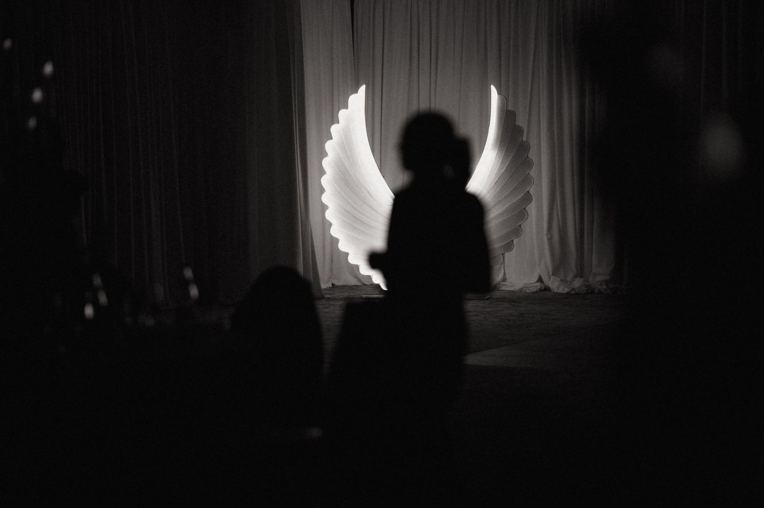 Silhouetted figure stands near a window with light streaming in, creating a dramatic contrast against dark interior.
