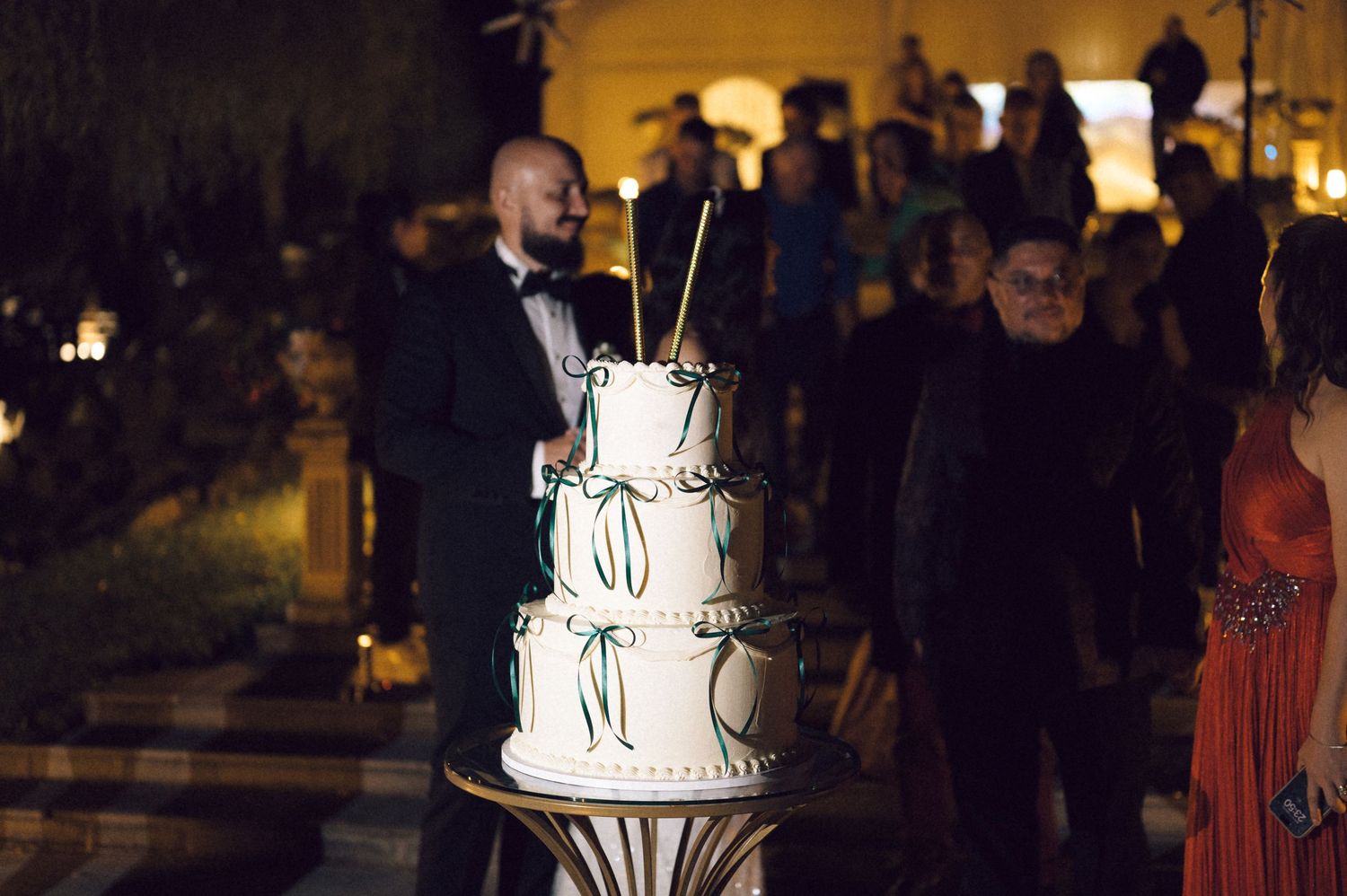 Wedding guests gather around an elegant white tiered cake at a dimly lit evening reception celebration.