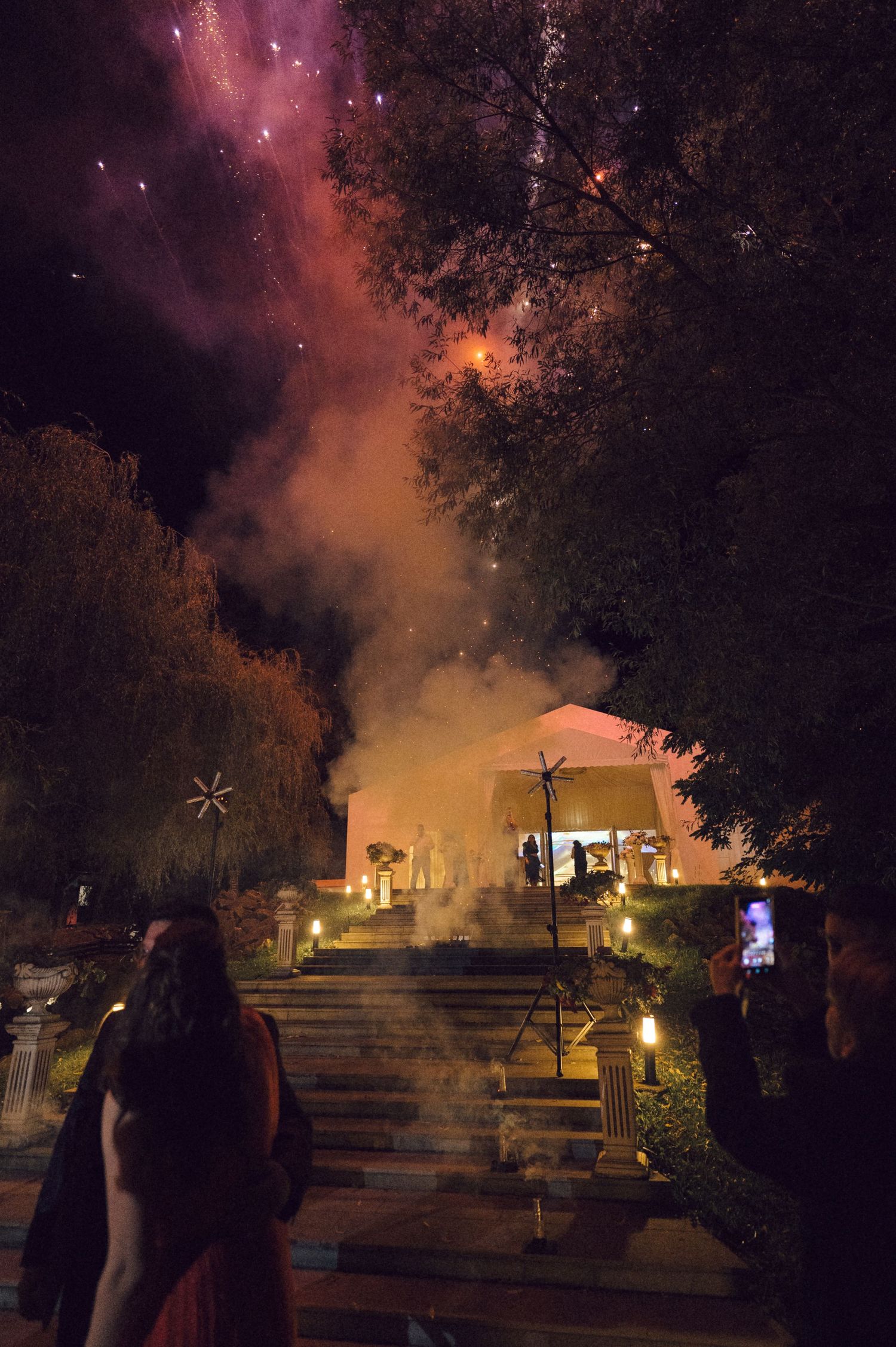 Nighttime view of illuminated buildings and steps through trees with misty orange and purple sky in the background.