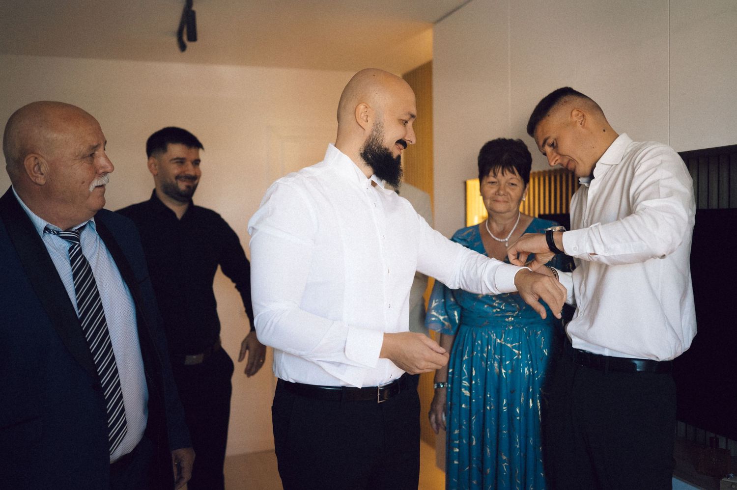 A group of people getting ready for a formal event, with someone adjusting a sleeve cuff in a bright interior space.