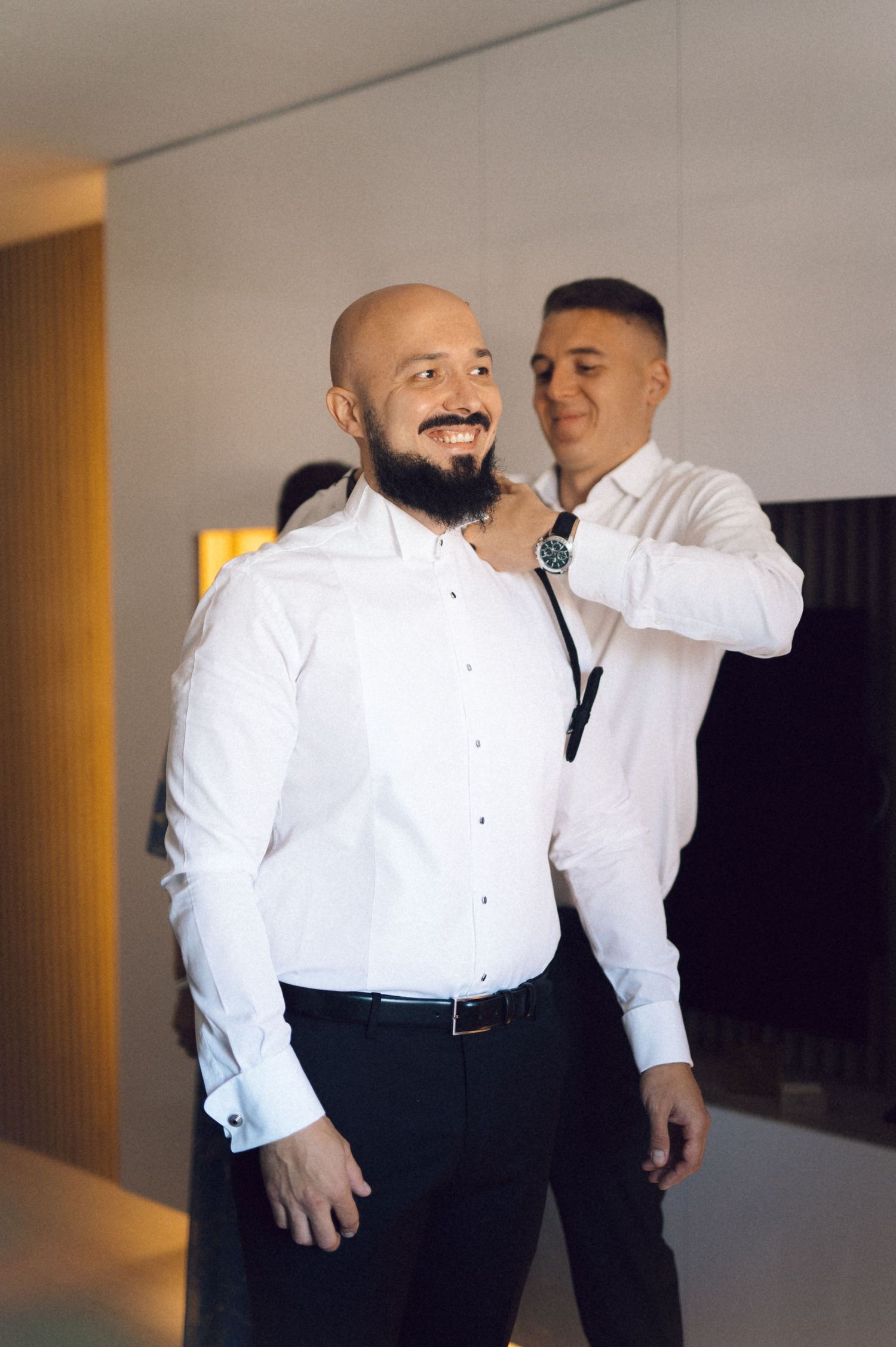 Two people in formal white dress shirts stand together in a hallway while getting ready for an event.