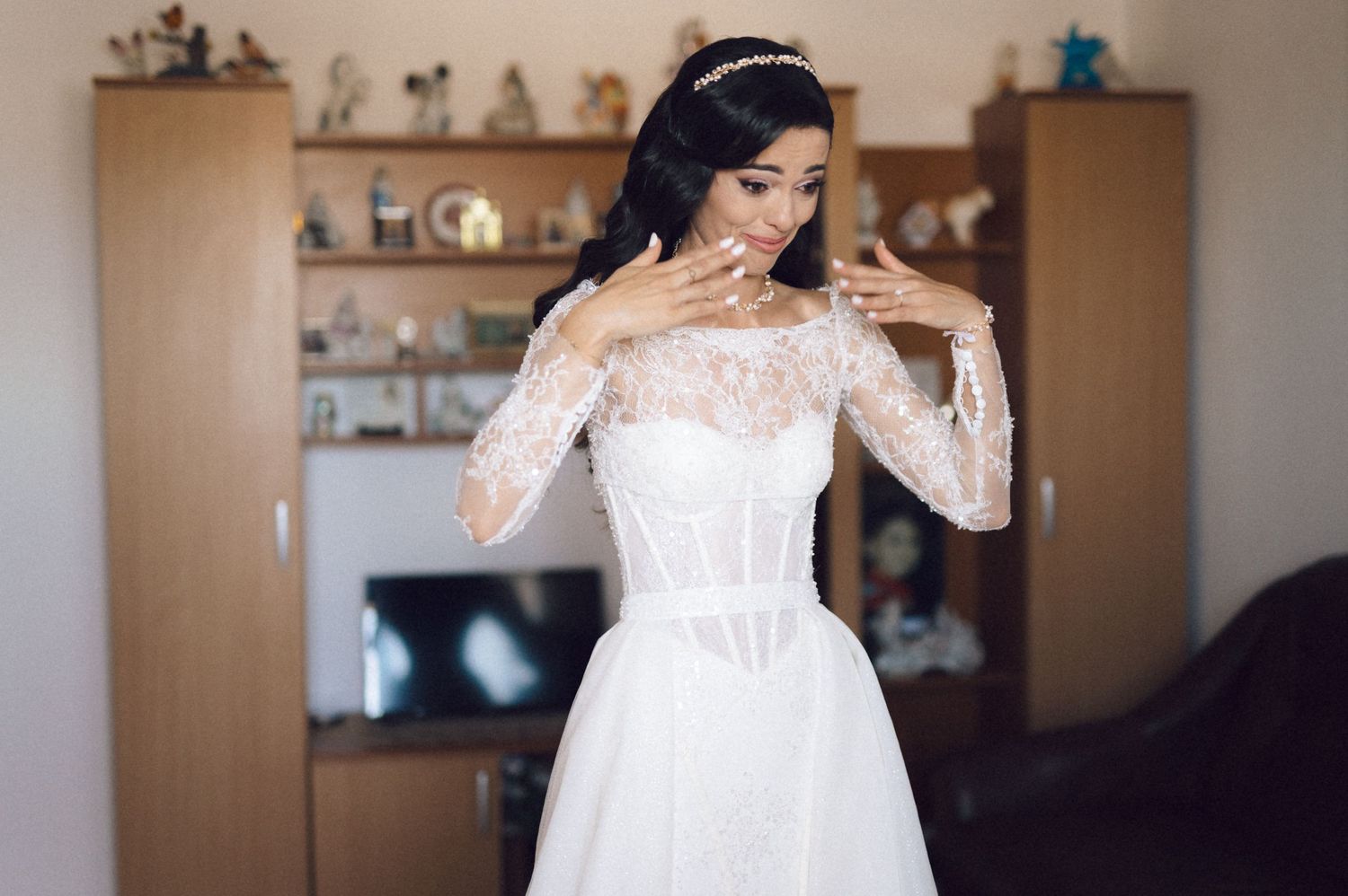 A bride in a white lace wedding dress laughs joyfully while getting ready in a room with shelves in the background.