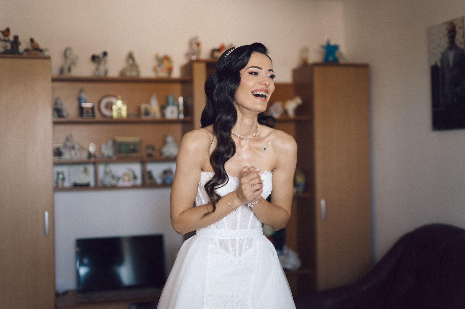 A bride in an elegant white gown stands in front of decorative wall shelves during wedding preparations.