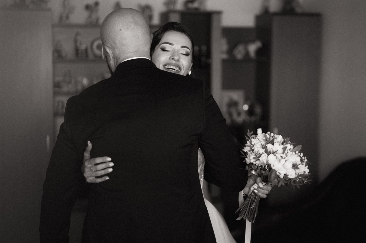 A couple shares a romantic dance together while holding a bouquet of white flowers in a dimly lit room.