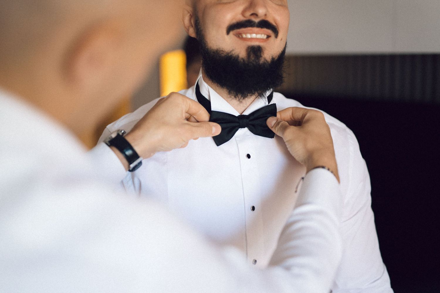 A person wearing a black bow tie and white dress shirt adjusts their collar in a formal setting.