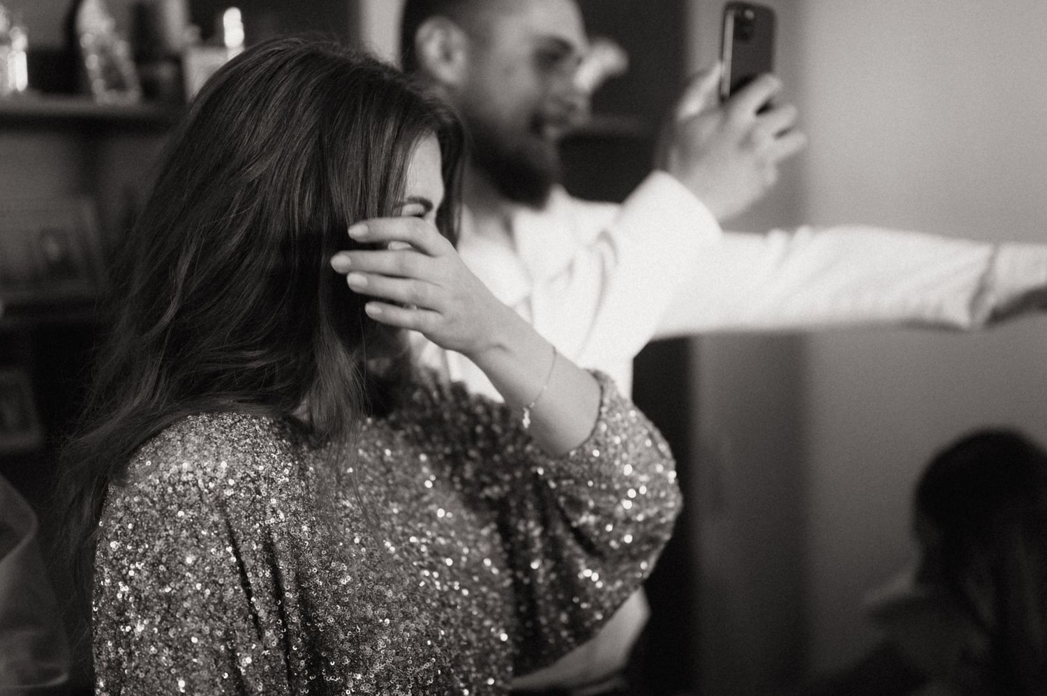 Black and white photo of someone in a sparkly dress covering their face while another person takes a selfie in background.
