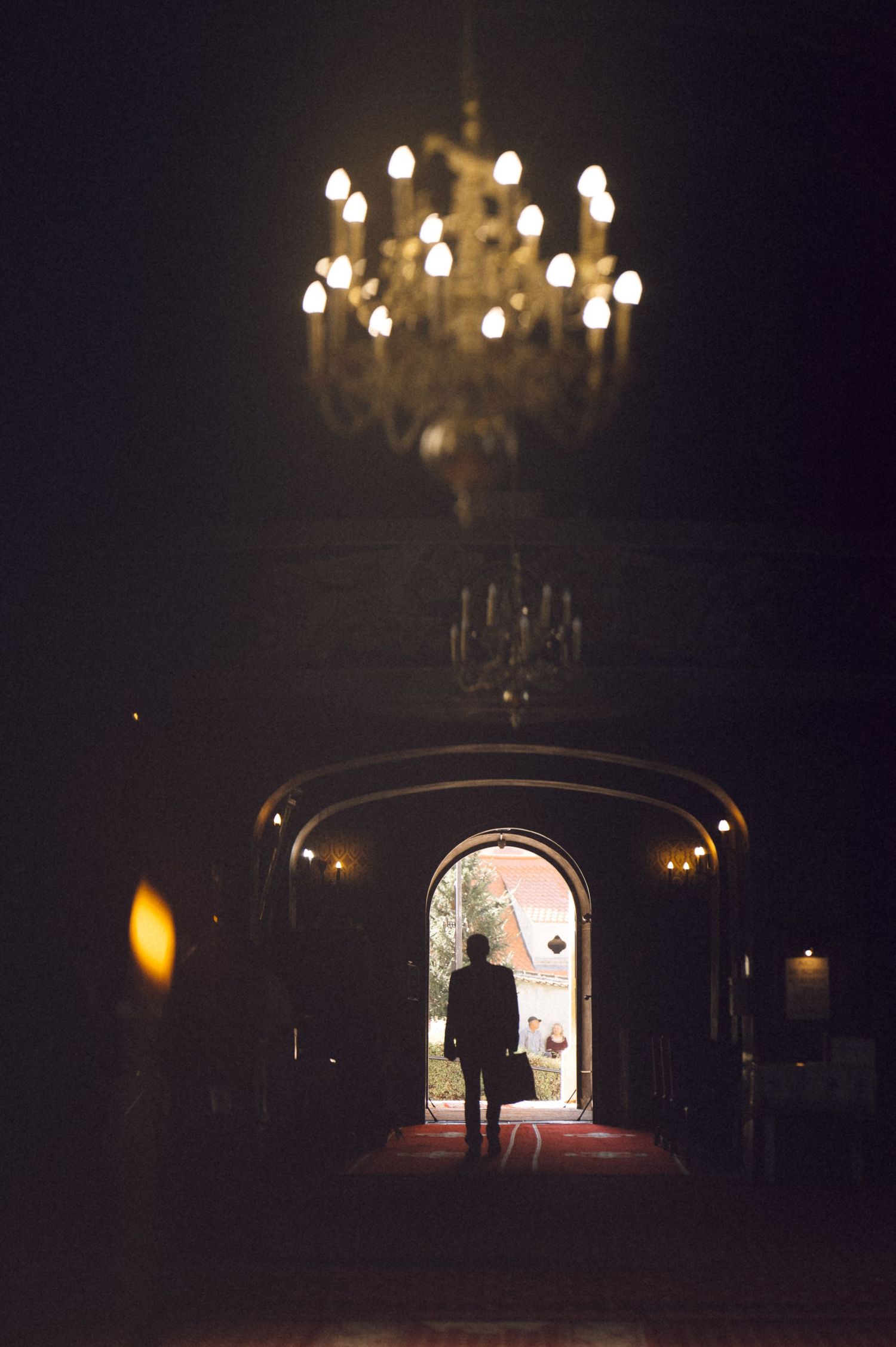 A silhouette of a person stands at the end of a dimly lit tunnel with chandeliers hanging overhead.
