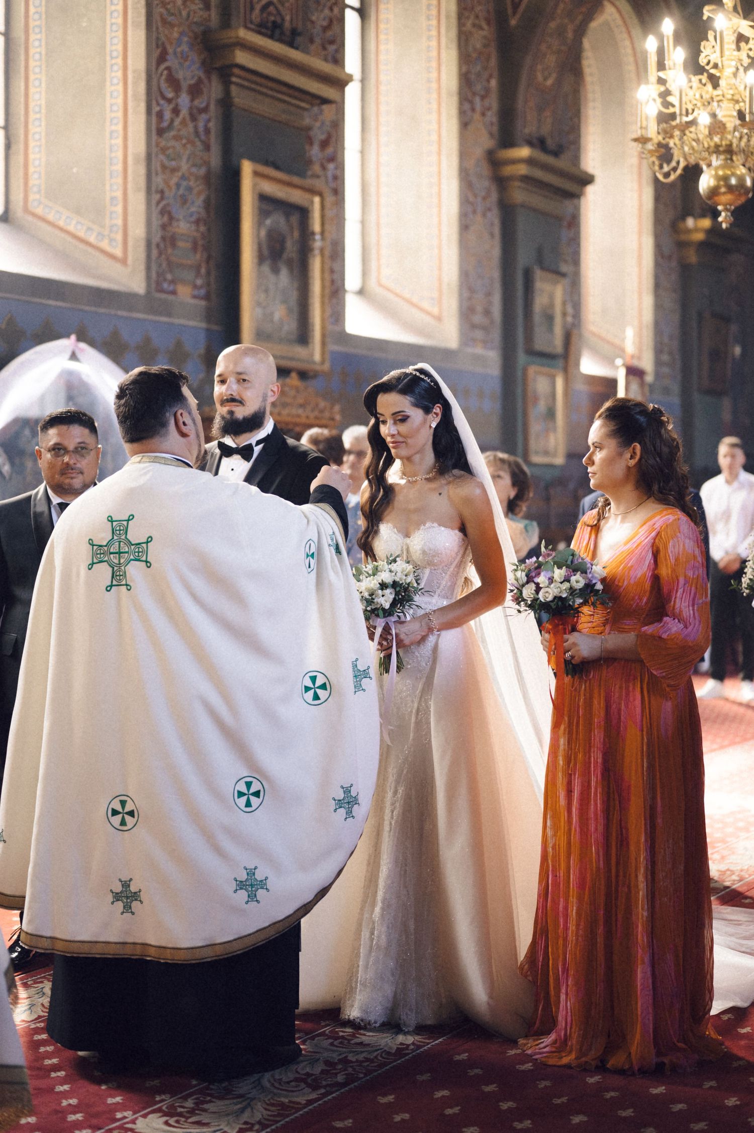A wedding ceremony takes place in an ornate church interior with a priest in white robes adorned with crosses.