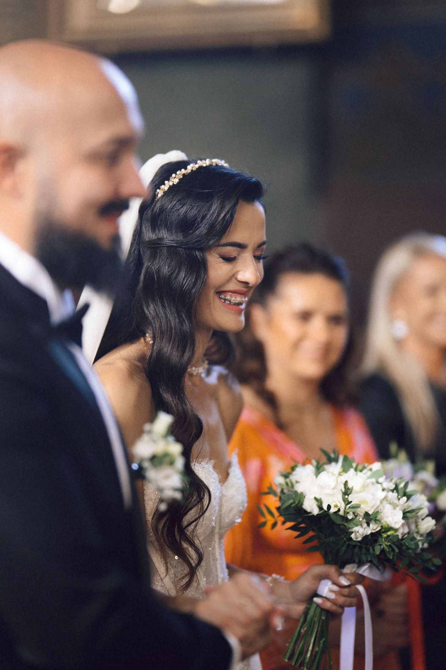 A bride in a white dress smiles while holding a white and green floral bouquet during a wedding ceremony.