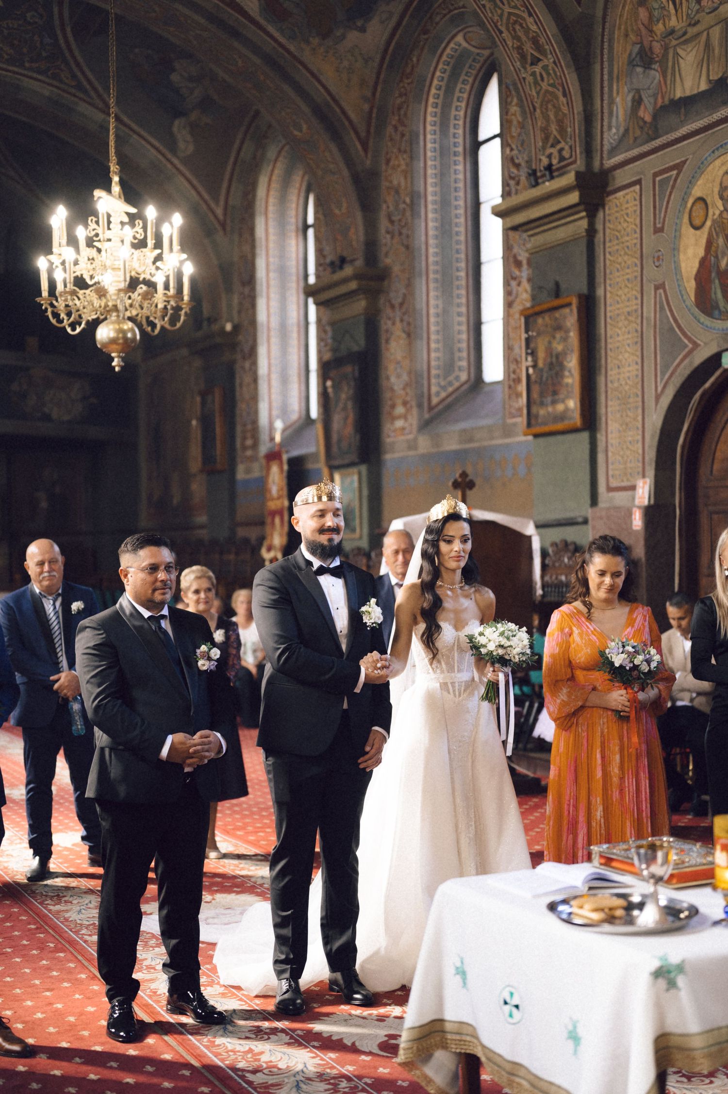 Guests gather inside an ornate Orthodox church during a religious ceremony with chandeliers and frescoed walls.