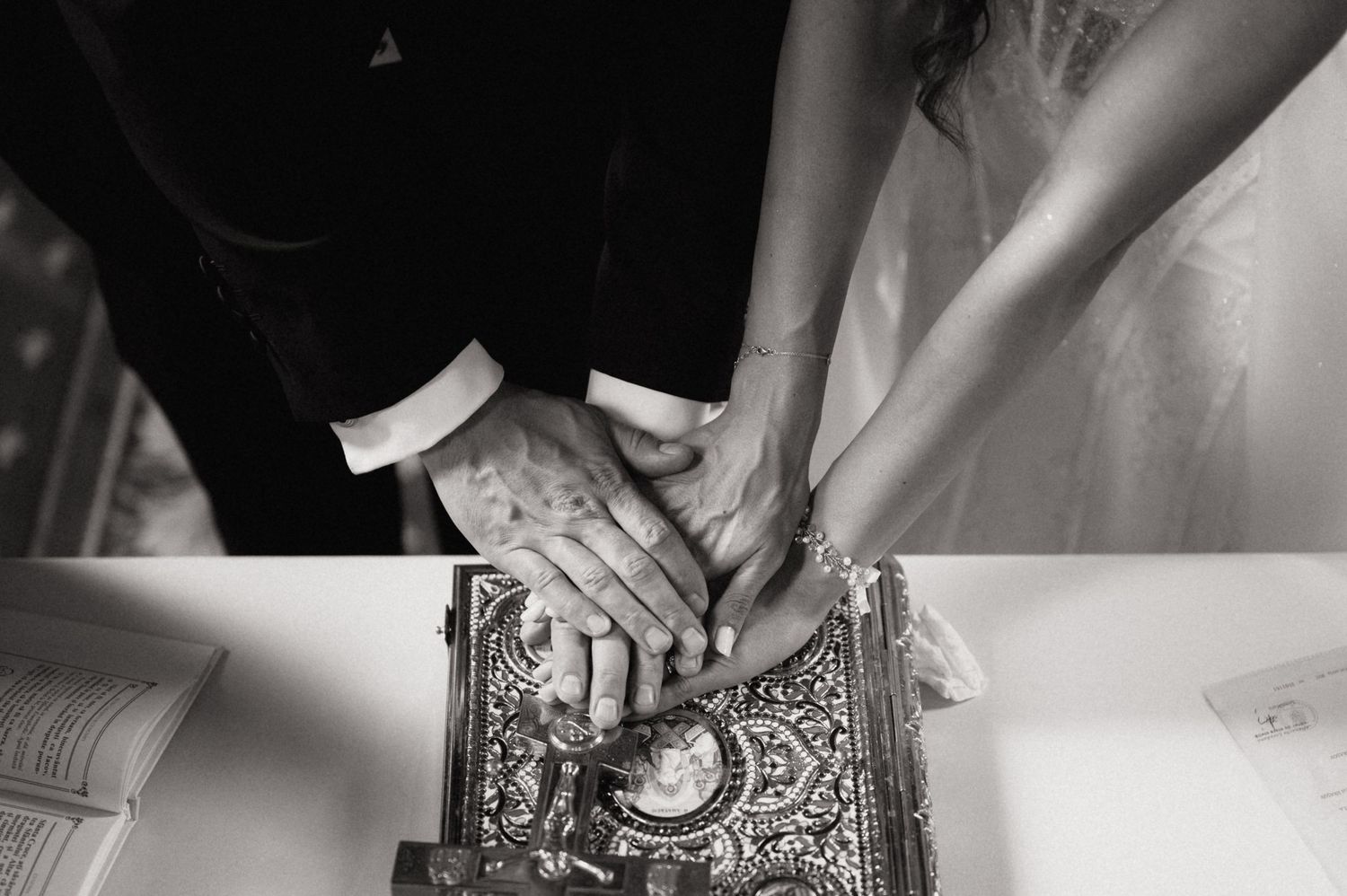 Two hands rest together on an ornate antique book during a wedding ceremony, captured in black and white.