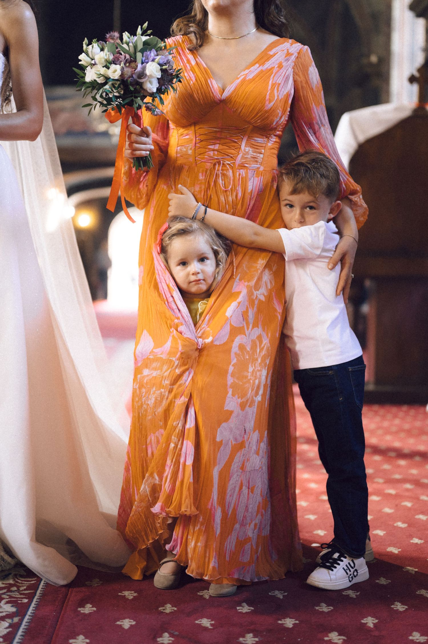 Small children peek out from behind an orange floral dress at a formal event with red carpeting.