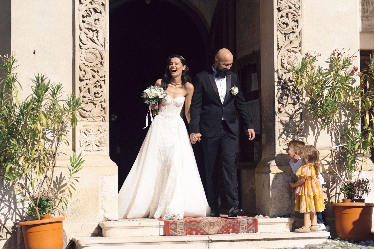 A couple in formal wedding attire stands in an outdoor courtyard with ornate stone columns and potted plants.