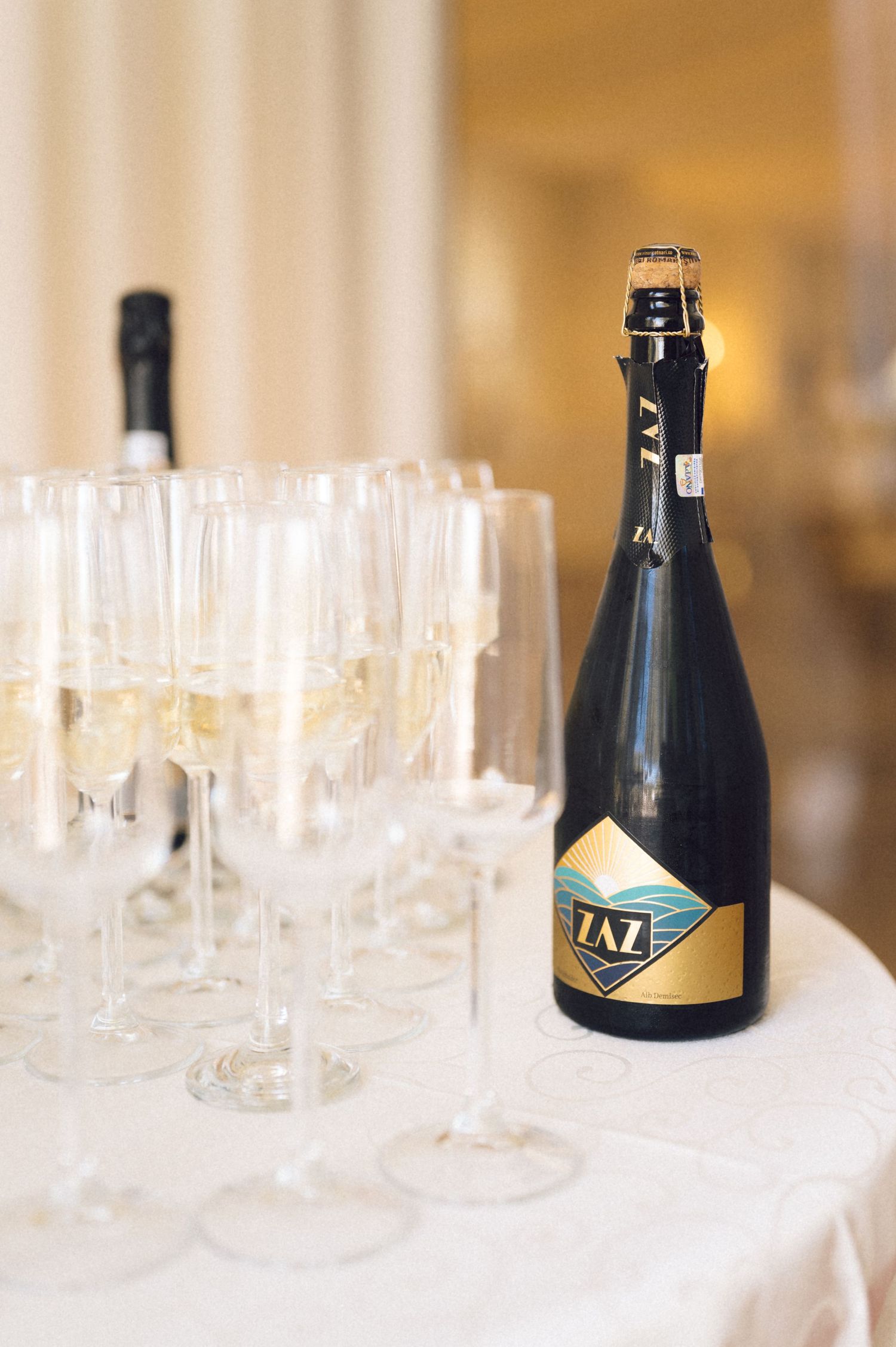 Empty wine glasses lined up on a white tablecloth with dark bottles in the background at a formal event setting.