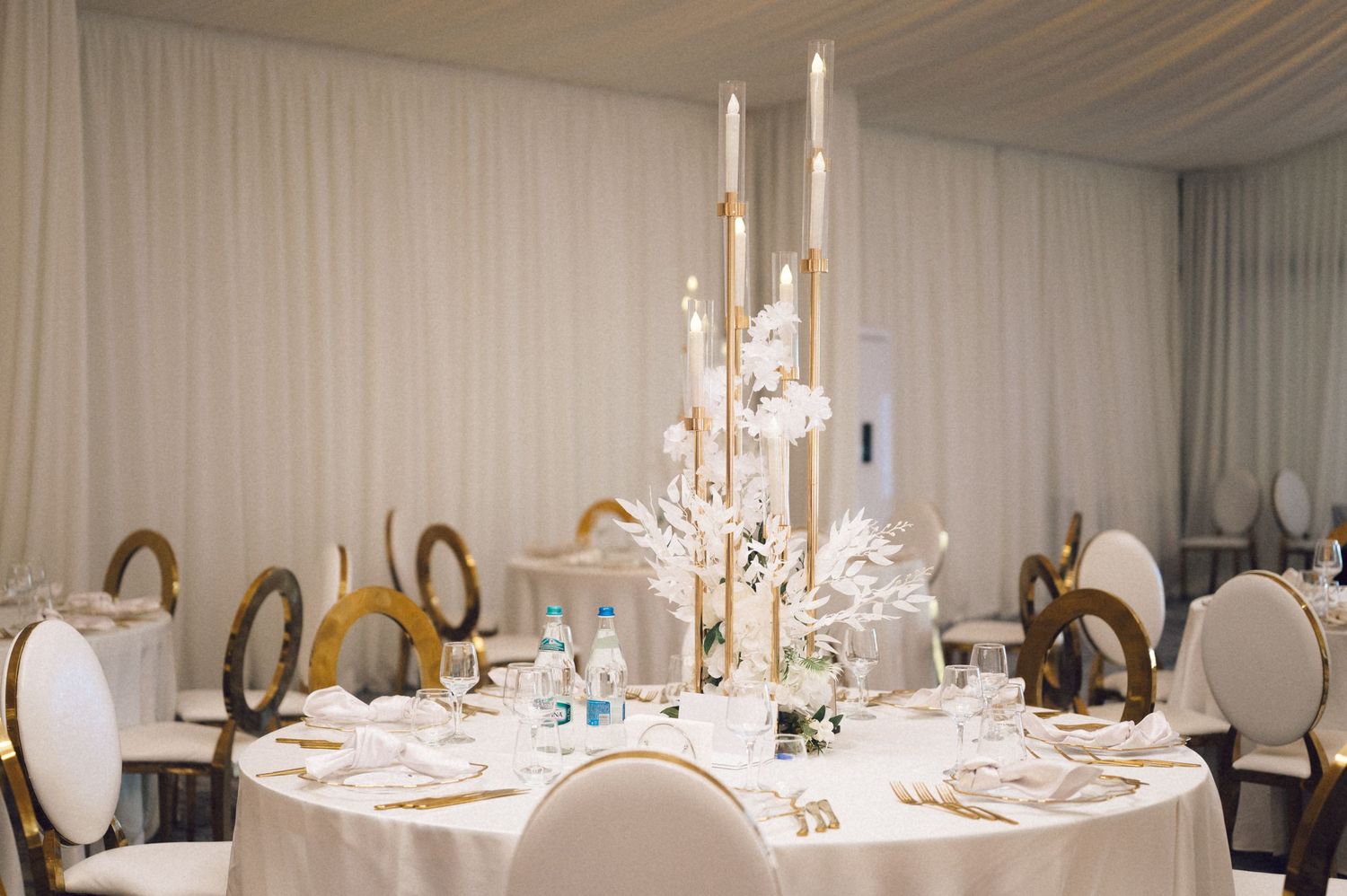Elegant wedding reception table with tall white floral centerpiece, taper candles and mixed white and gold chairs.