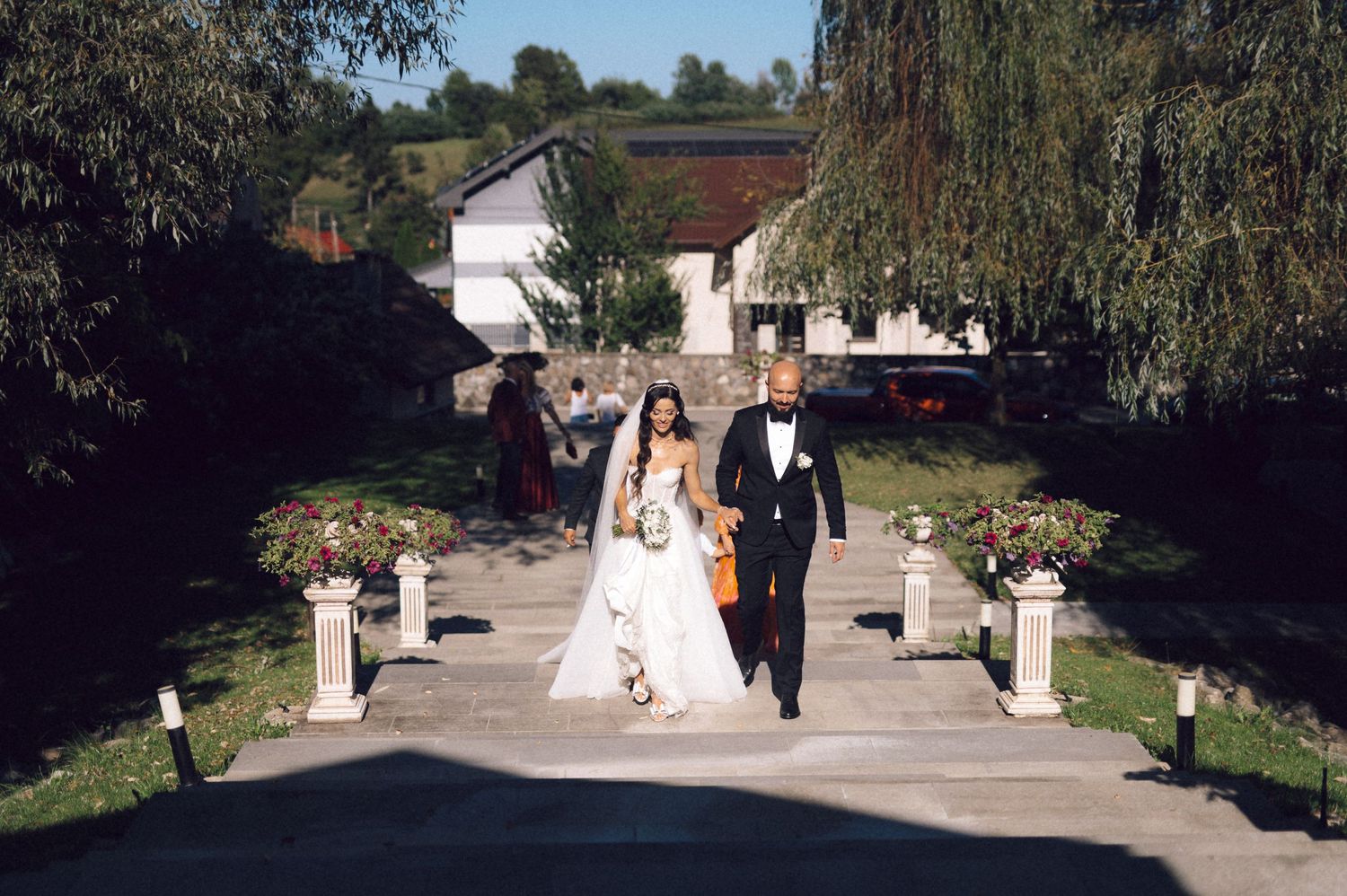 A couple in formal wedding attire walks down a tree-lined path during an outdoor ceremony.
