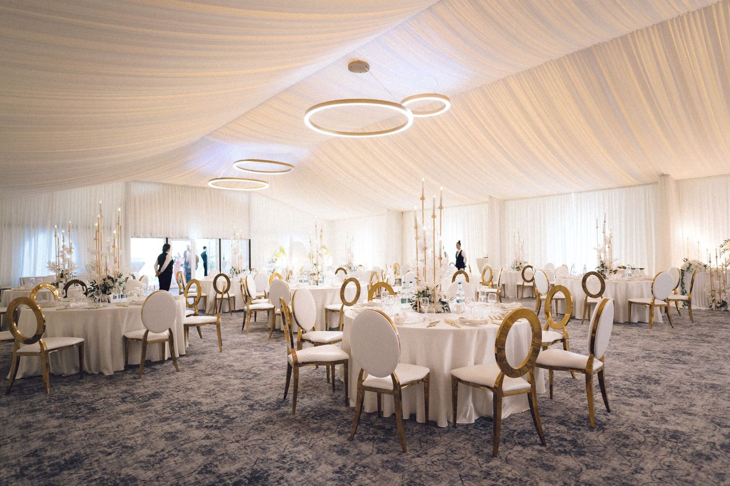 Elegant white draped wedding reception venue with round tables and ornate gold chairs set up for a formal celebration.