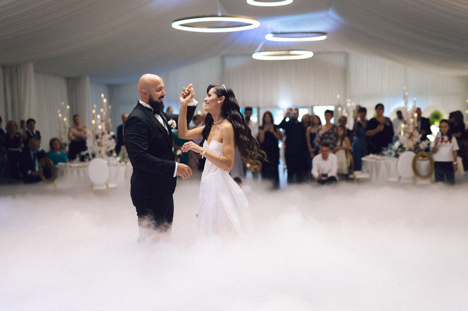 Wedding couple dances in a white reception hall filled with guests while fog covers the dance floor.