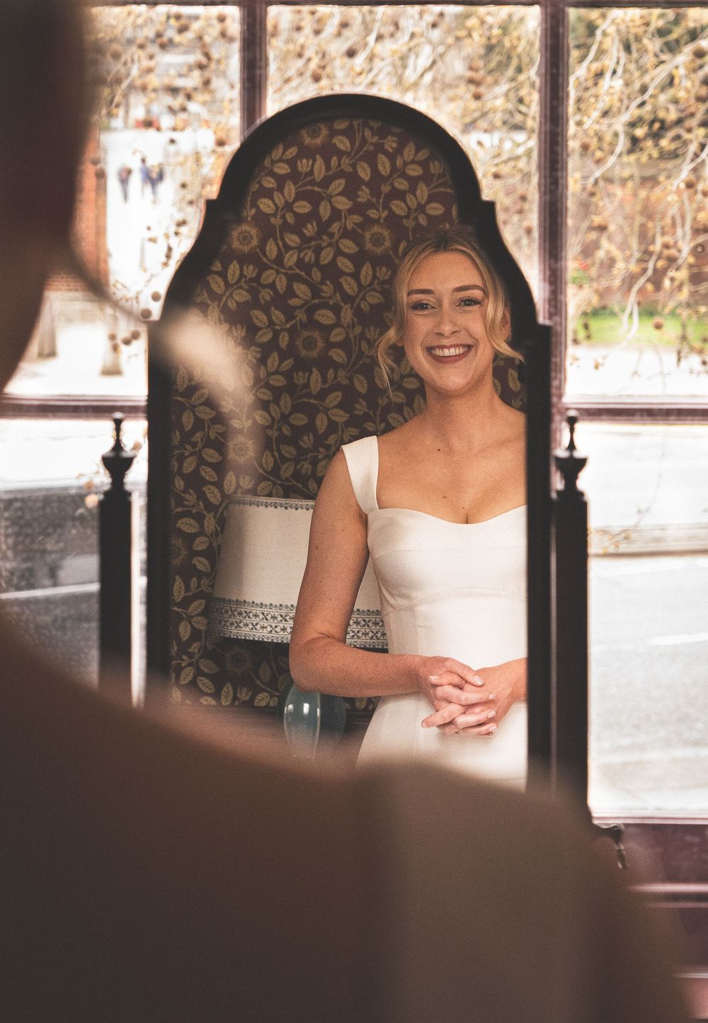 Smiling bride in white gown standing before ornate dark floral chair during wedding ceremony indoors.